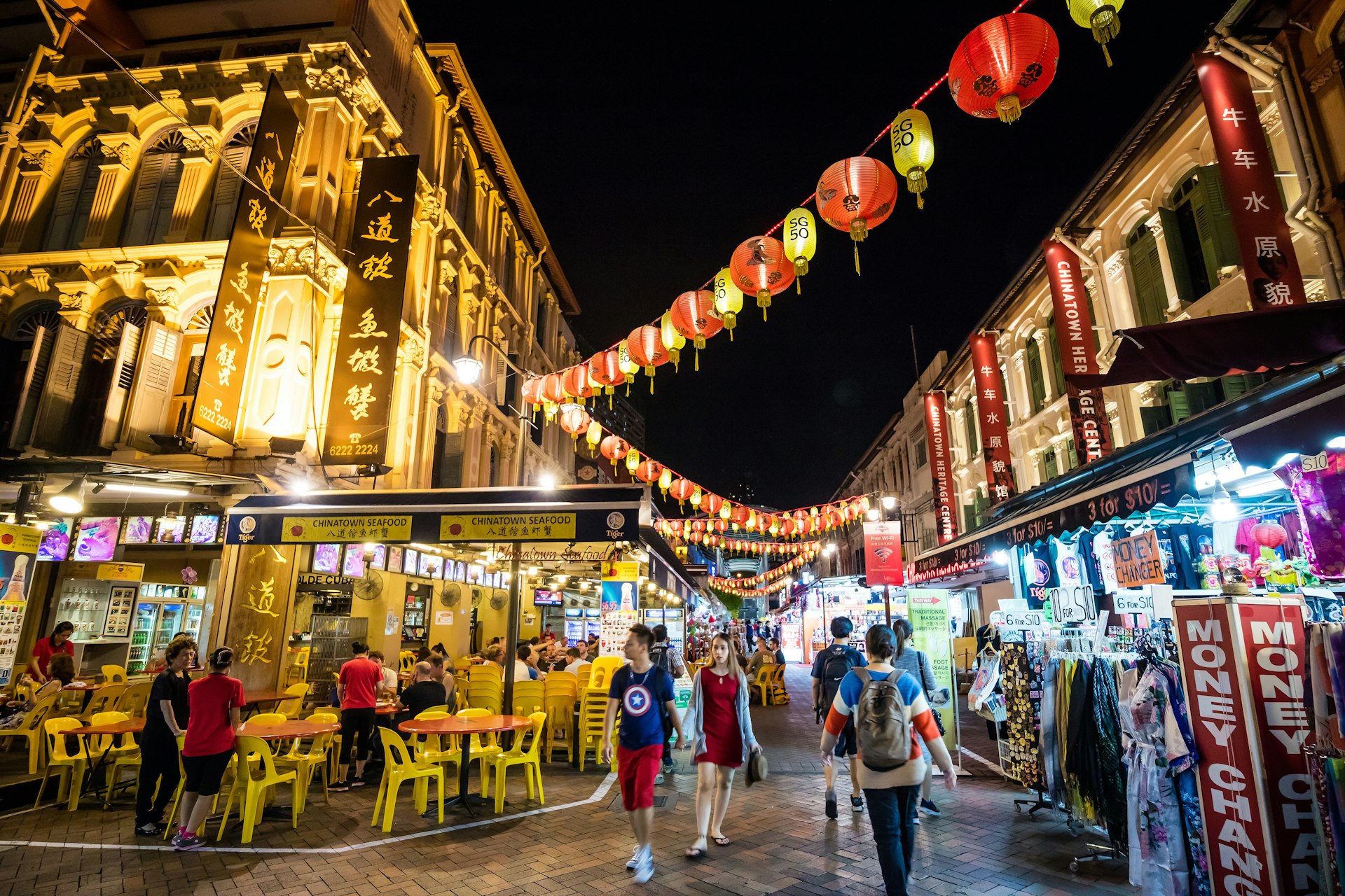 The Chinatown district in Singapore with diners sat on yellow plastic chairs and tables outside restaurants at night with lanterns hanging across the street lit up.