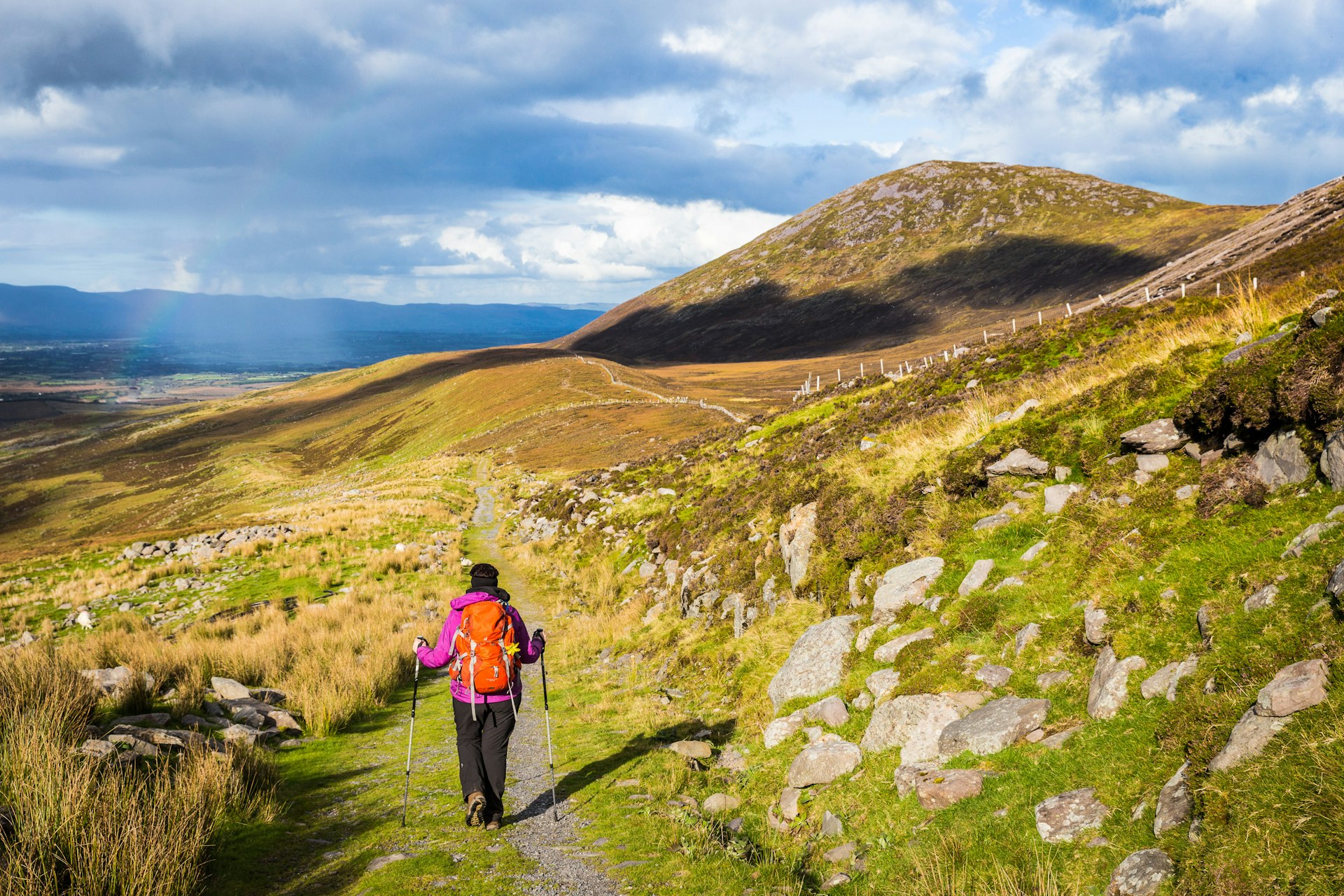 A hiker with a backpack and hiking poles follows a trail through a mountainous region of Ireland