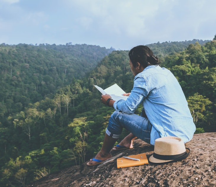 Asian man relaxing reading book on rocky cliff in mountains, Thailand