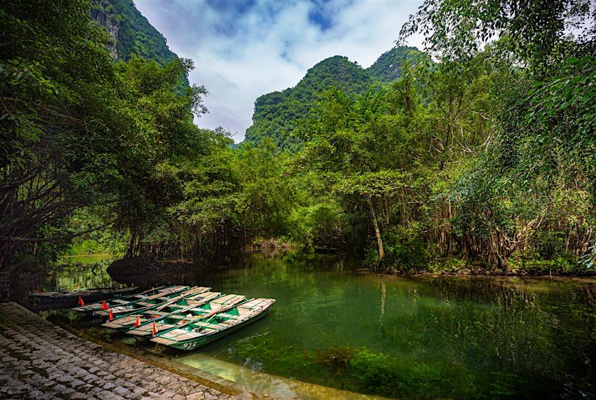 Small boats lining the shore of a river flanked by dense forest, with tall mountains behind