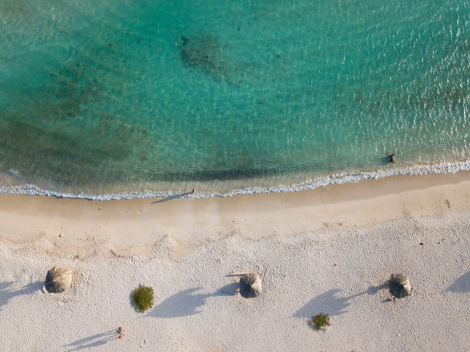 Aerial view of blue green waters at Baby Beach. On shore, you can see the roofs of three huts.