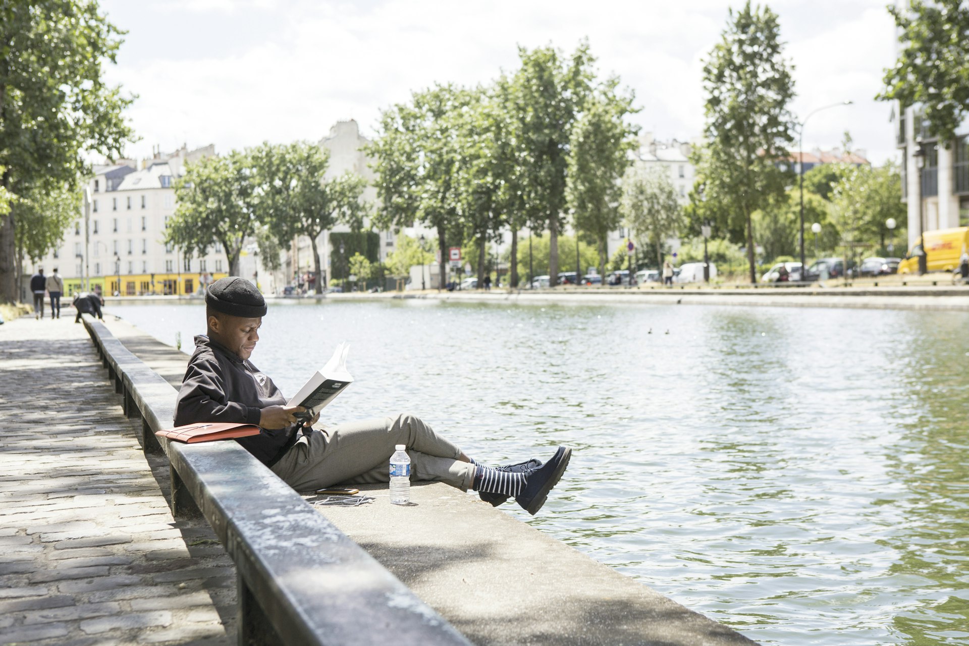 A man sits on the side of the Canal St-Martin in Paris reading a book on a sunny day.