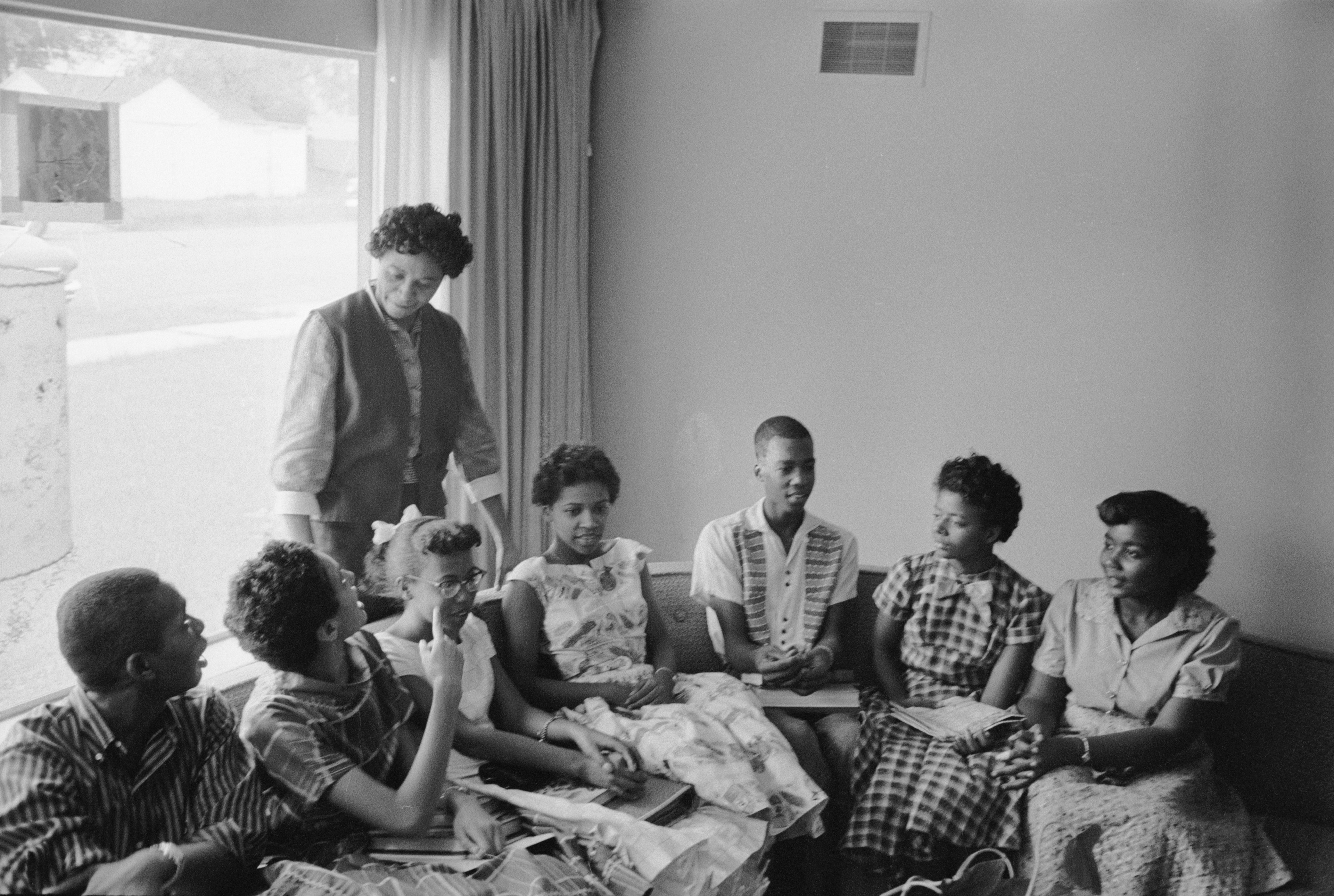 A woman stands behind a group of teenagers sat on a sofa: American Civil Rights leader & journalist Daisy Bates (standing) talks with some of the Little Rock Nine at her home in Little Rock, Arkansas October 1957.