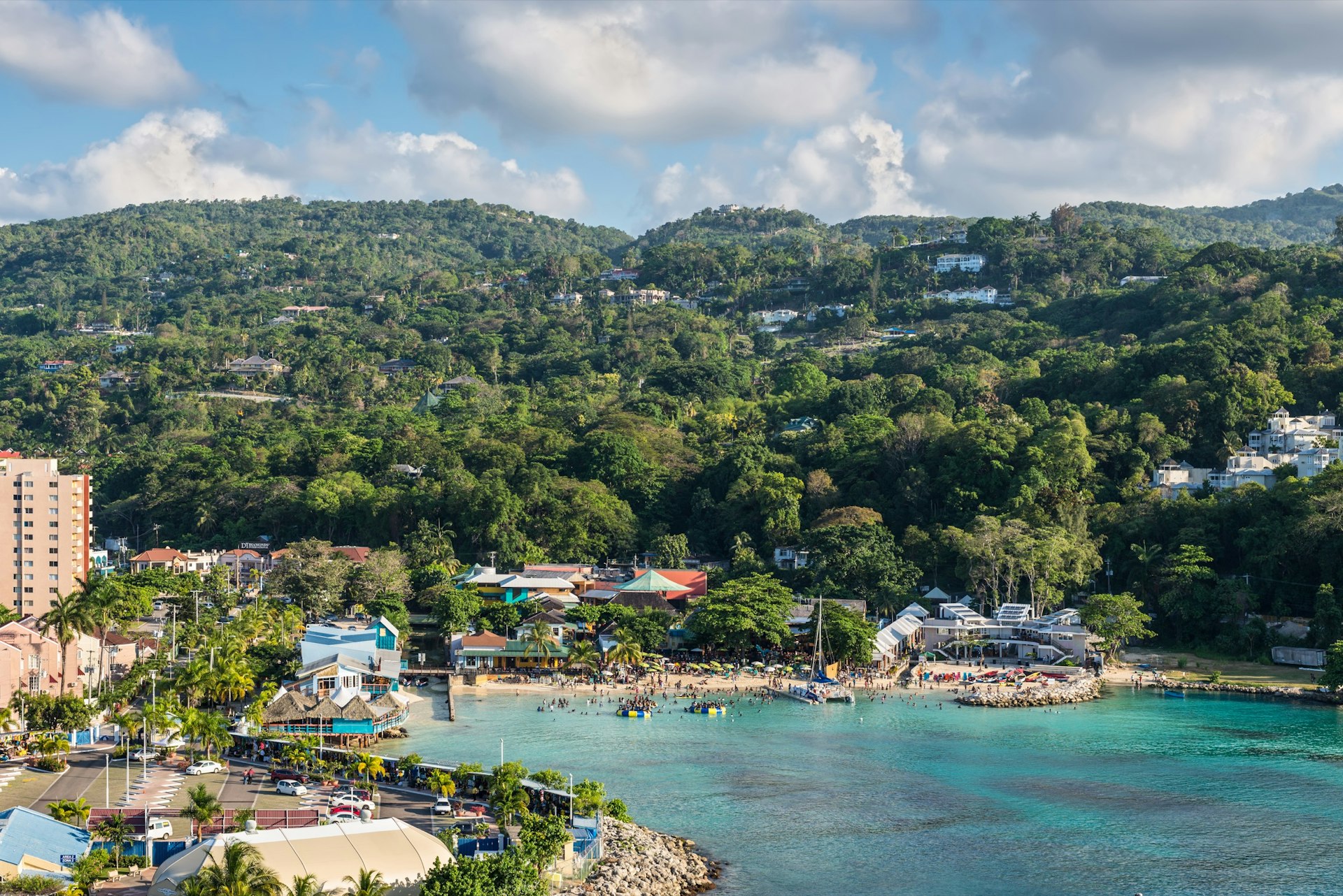 View from a ship of the blue waters of Fisherman's Beach in Ocho Rios, Jamaica. There are large groups of people are on the beach, there are large inflatable structures in the ocean and a sailboat docked in the shallow waters. 