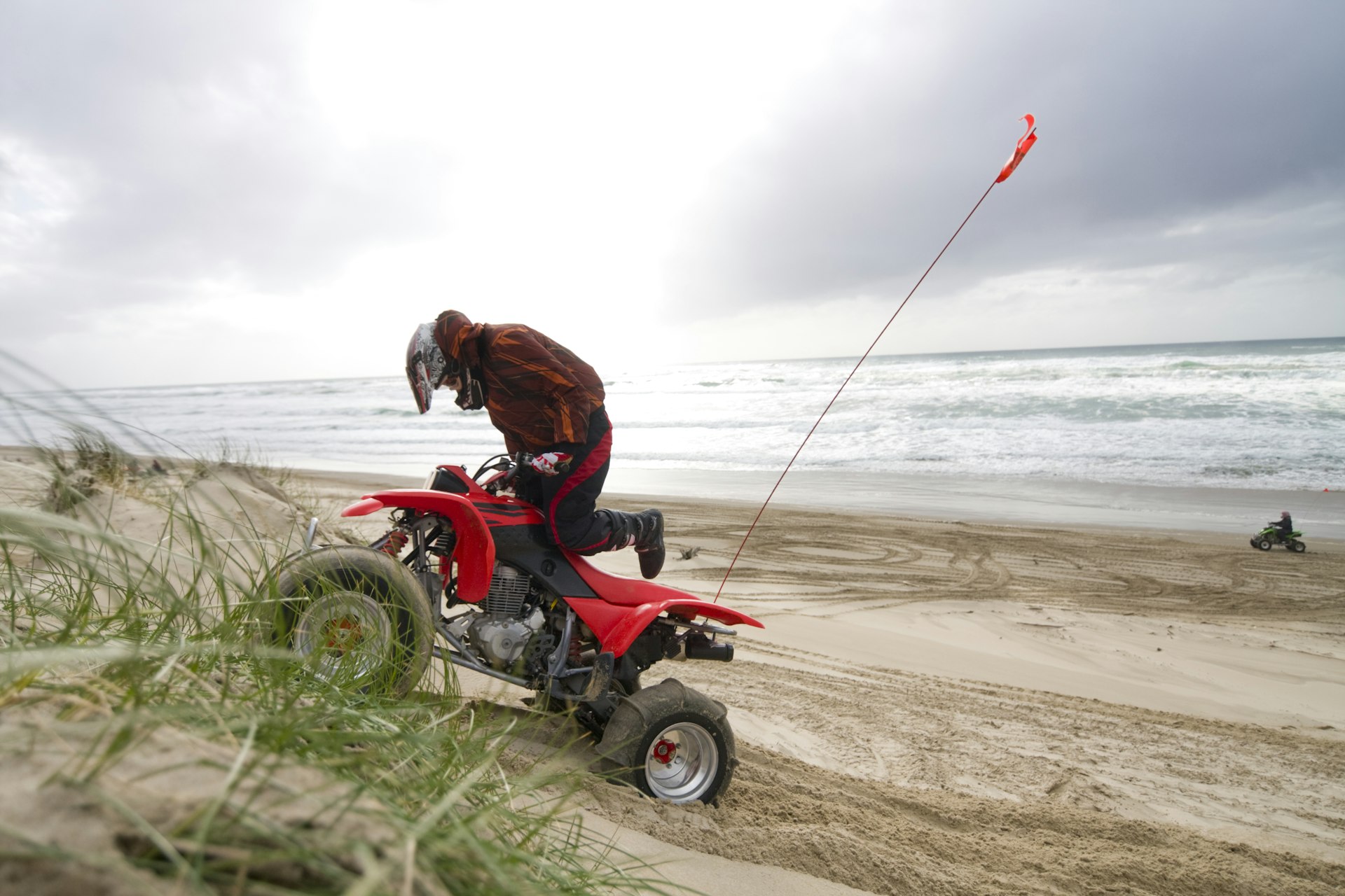 Quad rider driving an ATV up a grassy hill on the Oregon coast beach. Second quad rider off in the distance closer to the ocean waves. 