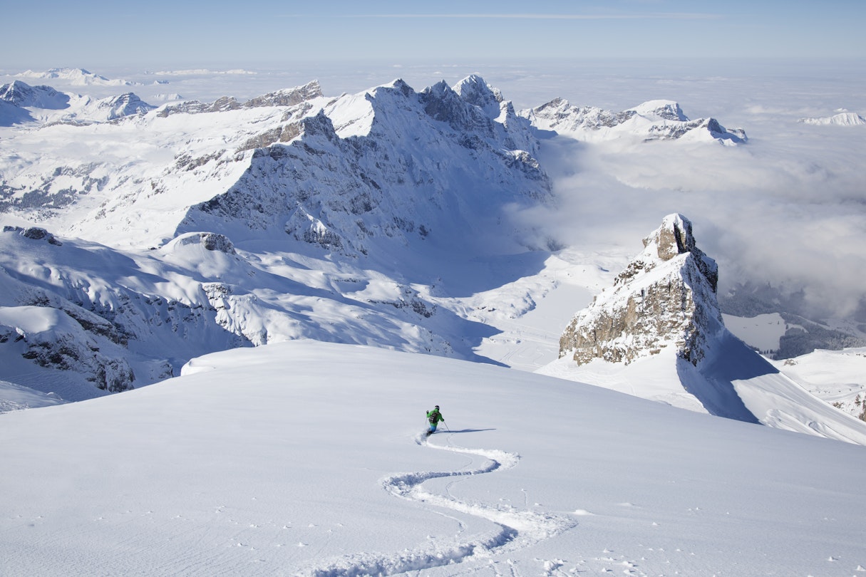 Off-piste skier carving lines through powder snow with the peaks of the alpes beyond.