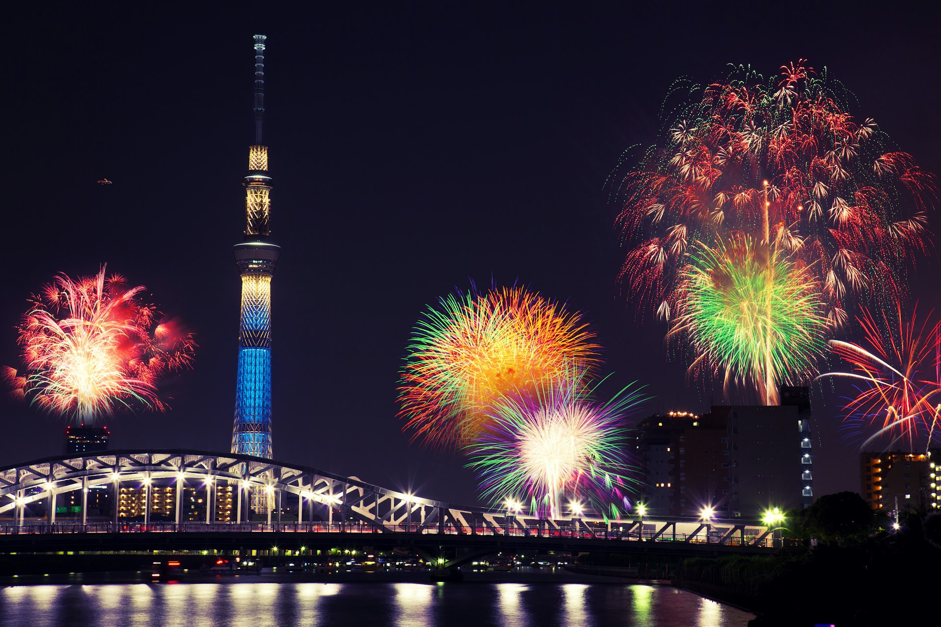 Summer Firework display over the Sumida River in Tokyo. Colourful explosions fill the night sky, below which a handful of skyscrapers are visible.