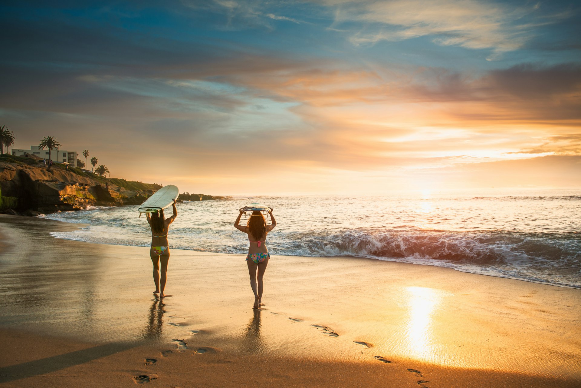 Two female surfers carry surf boards, walking along a beach in La Jolla, San Diego at sunset