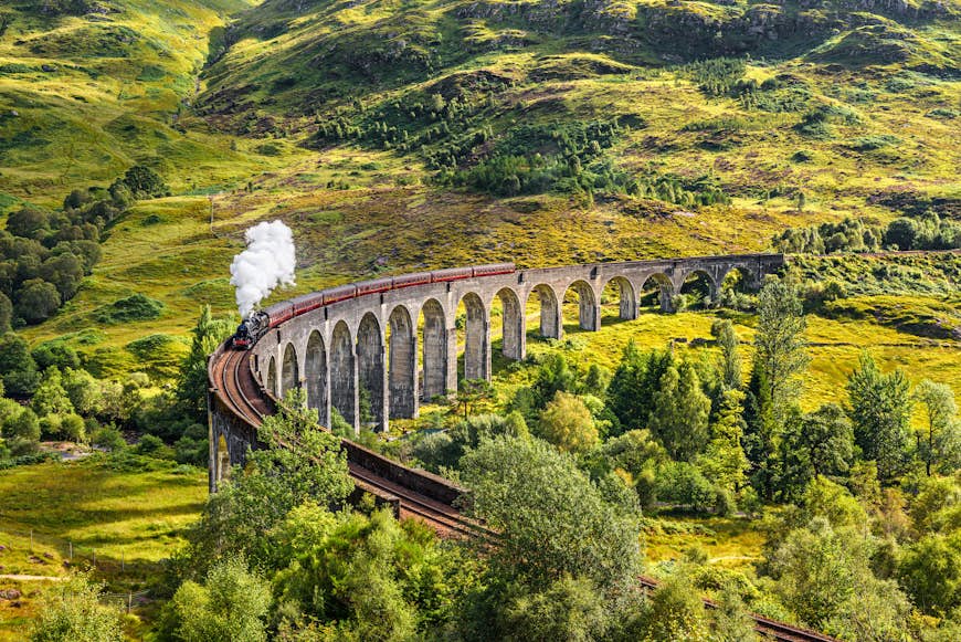 A viaduct with many arches rises above green hillsides. A steam train chugs along it.