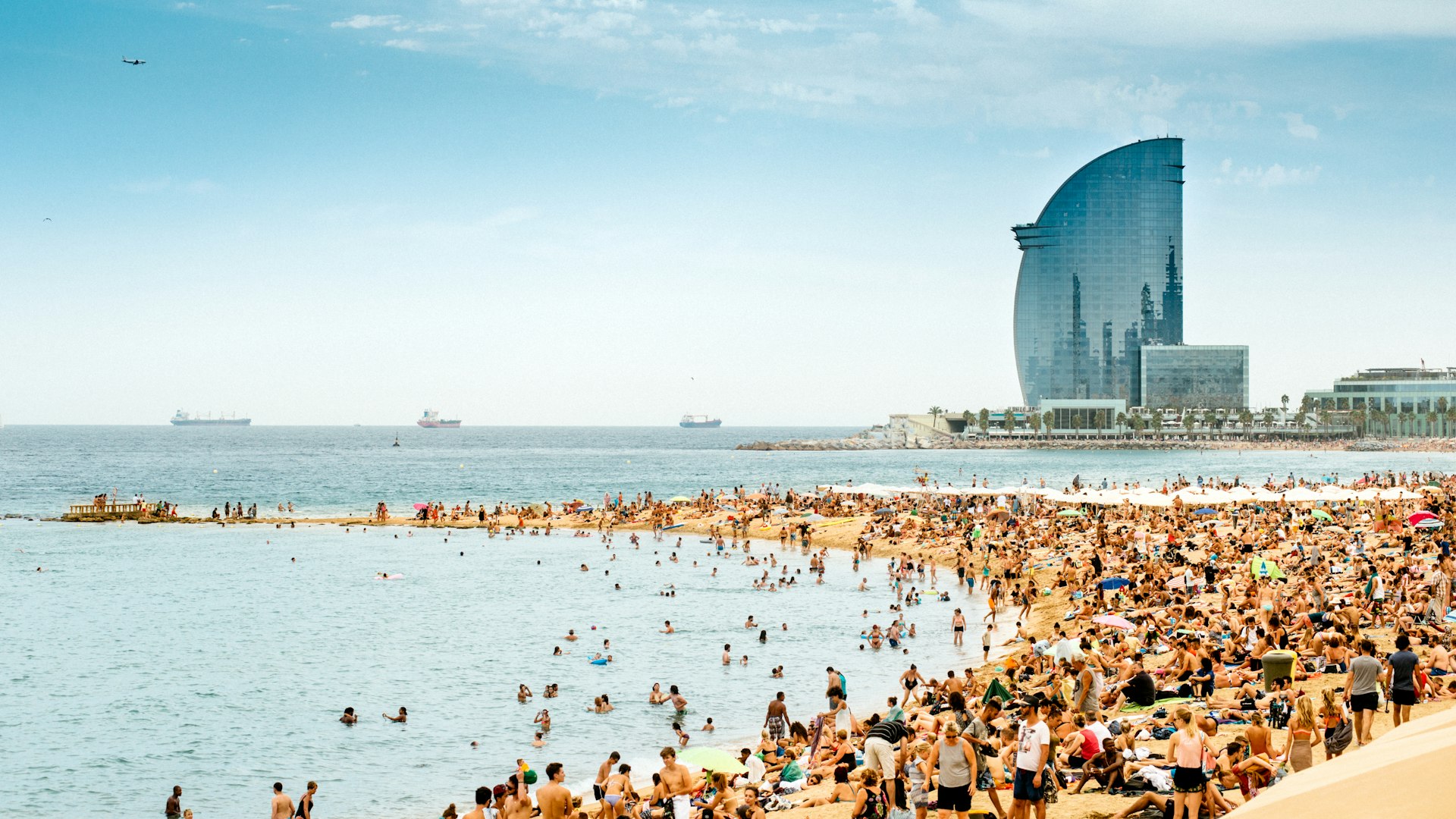 A crowded beach full of people enjoying the sunshine. A curved steel-and-glass building is at the end of the beach