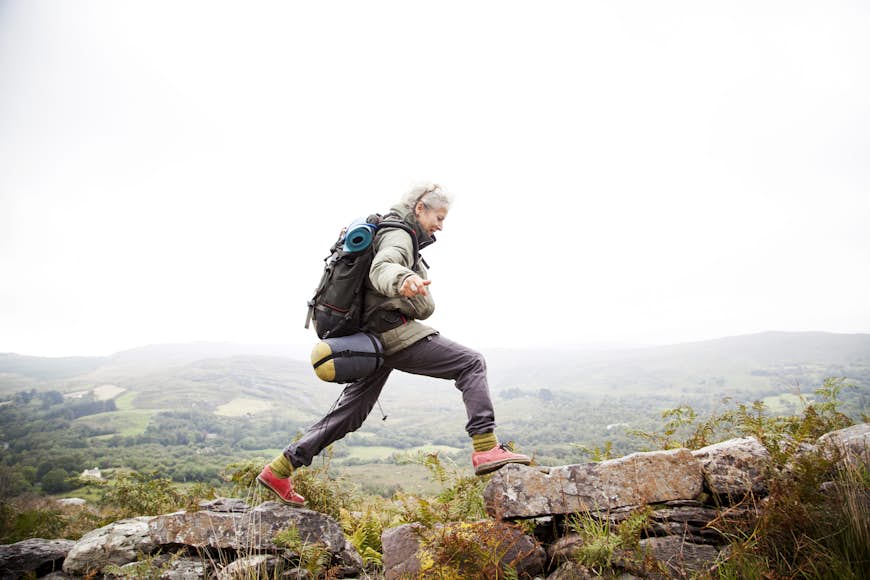 An older woman jumps across rocsk in the mountains of Ireland