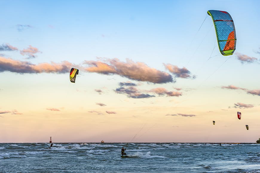 Kitesurfers out at sea with large colorful kites flying above them against an orange sky
