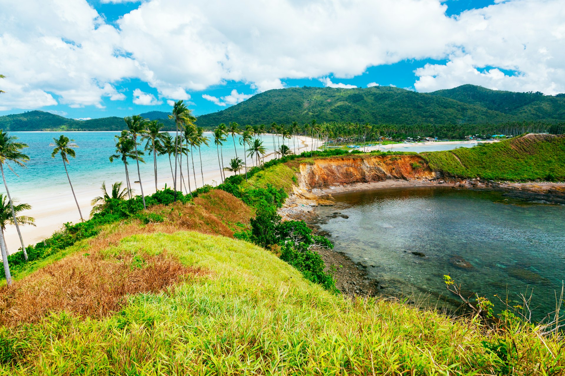 An aerial view of Nacpan Beach near El Nido in the Philippines. The white-sand beach is backed by palm trees and has a lake directly behind it.