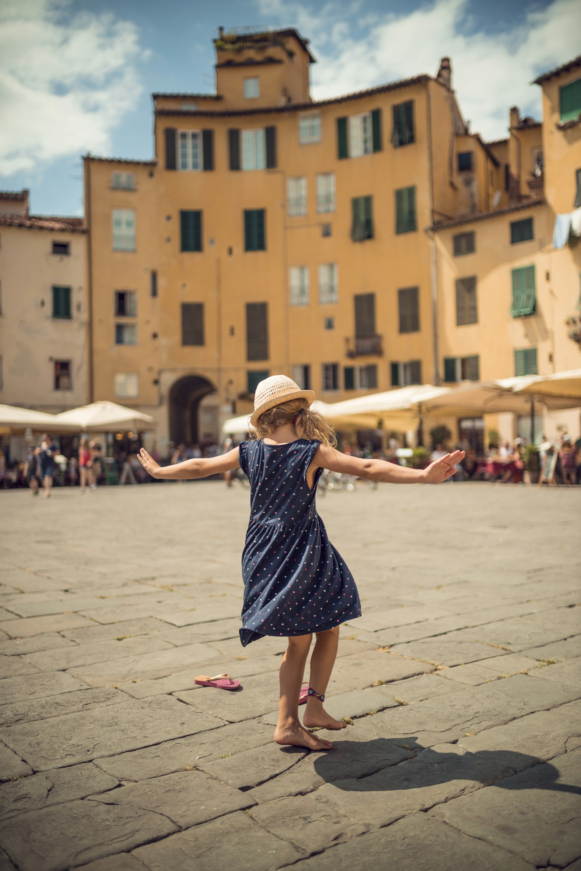 Back view of little girl dancing in a large public square surrounded by Renaissance buildings, and lined with umbrellas