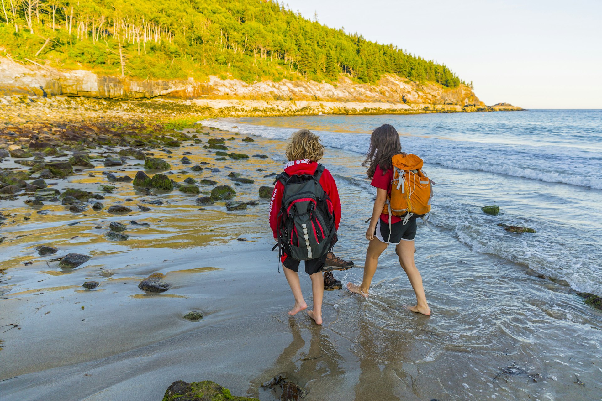 A teenage boy and girl carrying backpacks walk barefoot on a sandy beach 