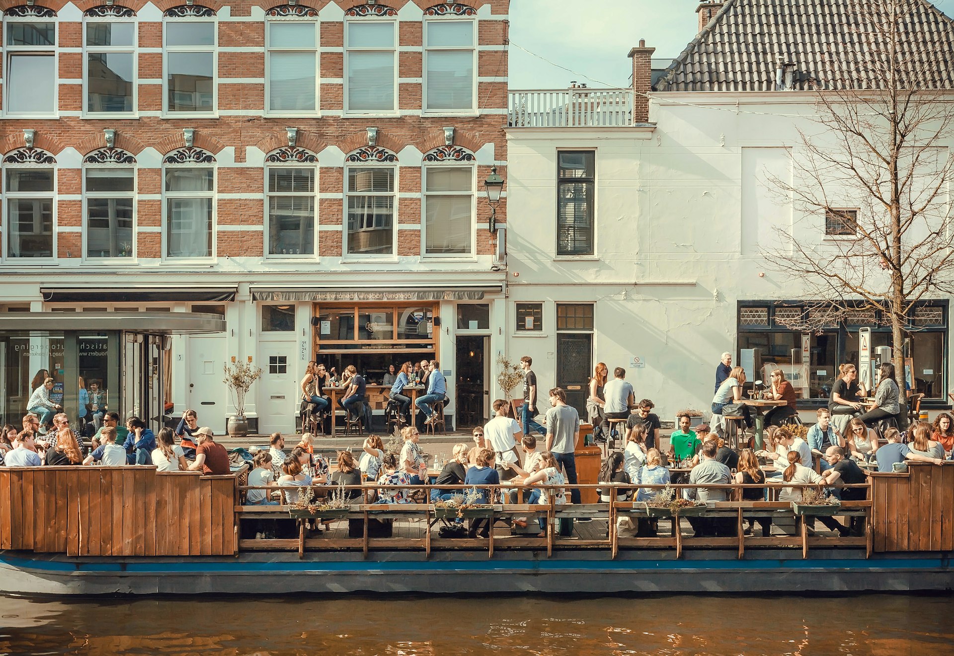 Crowds of people eat and drink outside Grapes and Olives riverboat cafe on the side of the canals in The Hague, the Netherlands