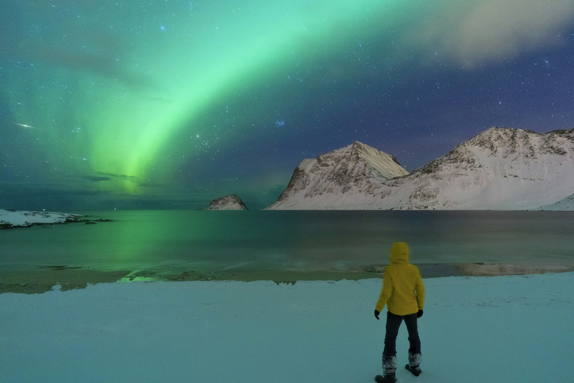 A person stands on a shore as greens and blues of the northern lights swirl in the sky above