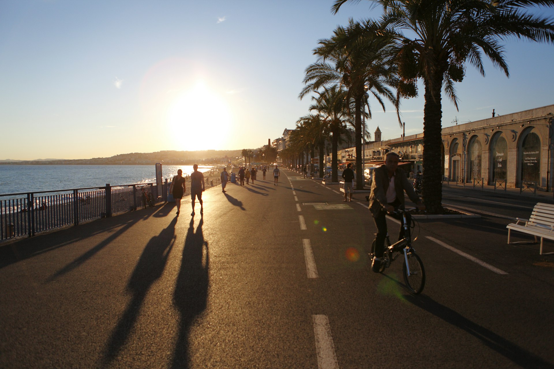 People walking and cycling seen in silhouette as the setting sun casts shadows on the Promenade des Anglais, Nice, Côte d’Azur, France
