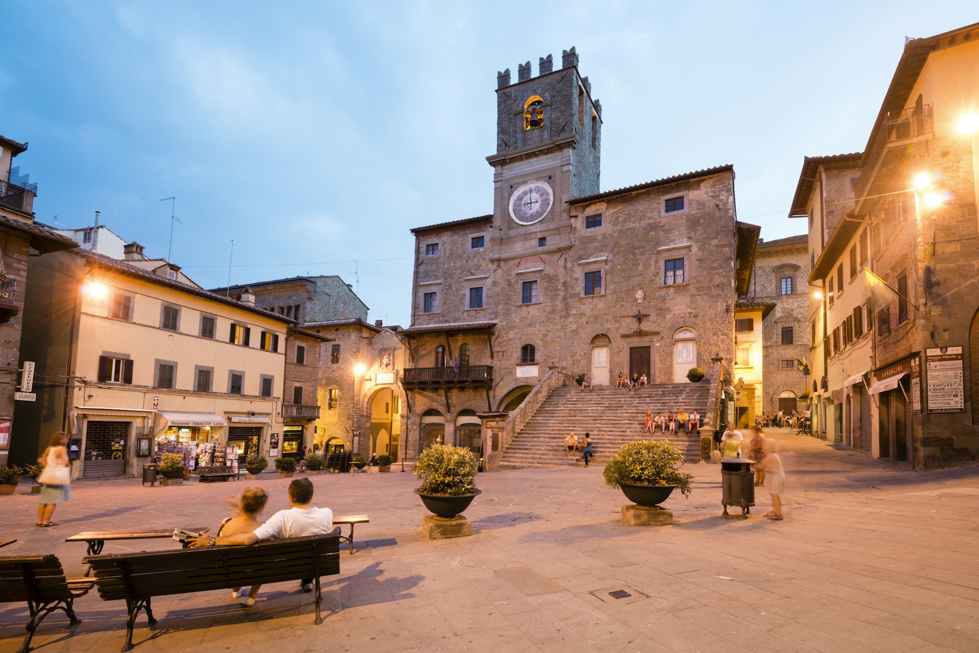 A medieval square at dusk. Two people sit on a bench and admire the surrounding architecture