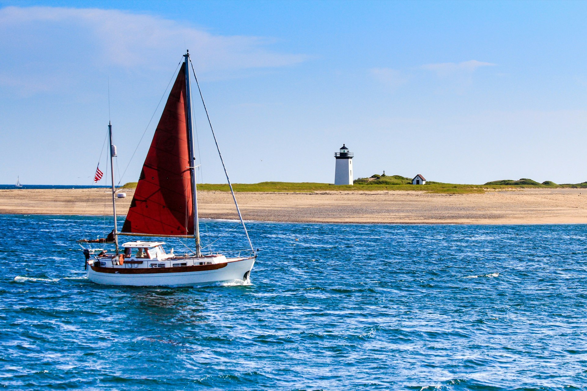 A sailboat with red sail sails past a white lighthouse and a little house with a red roof on land at Long Point Beach.
