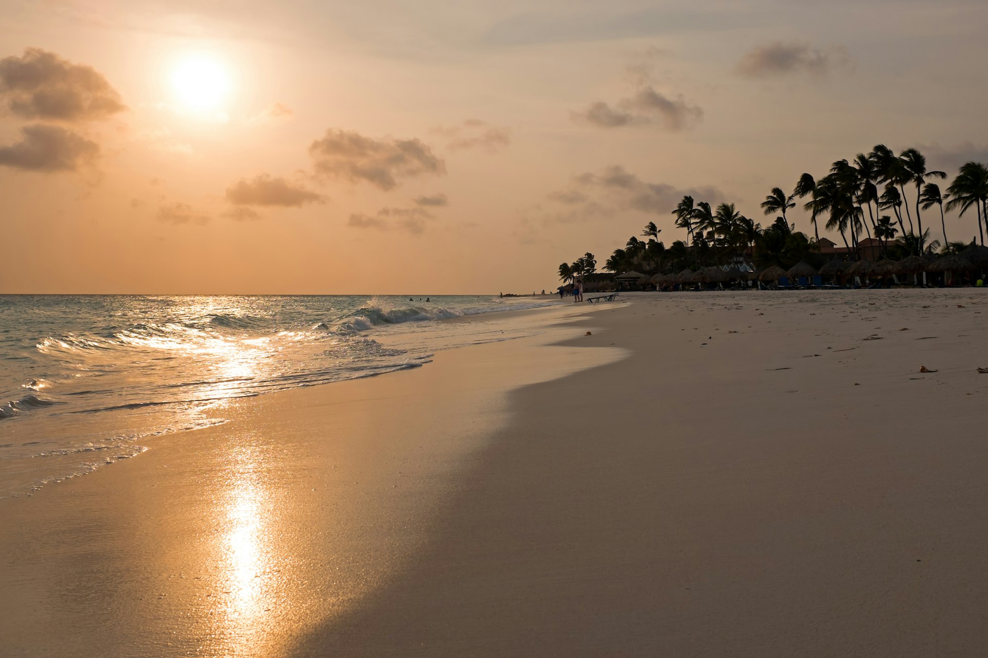 The sun sets on a sparely populated beach in Aruba. There are tall palm trees swaying in the wind in the background.