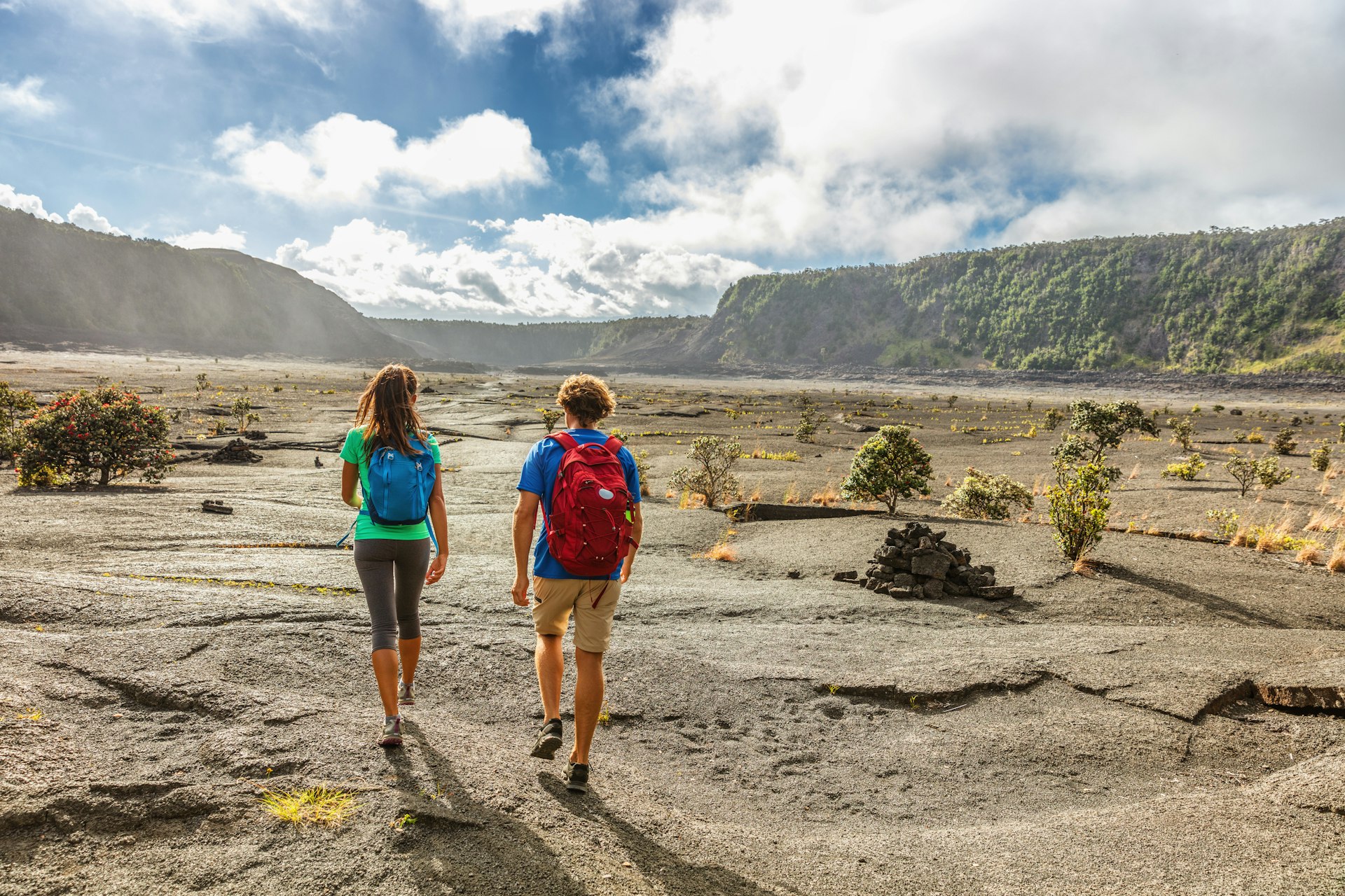 Two hikers walk away from the camera on a hiking trail across a crater, a large dusty expanse 