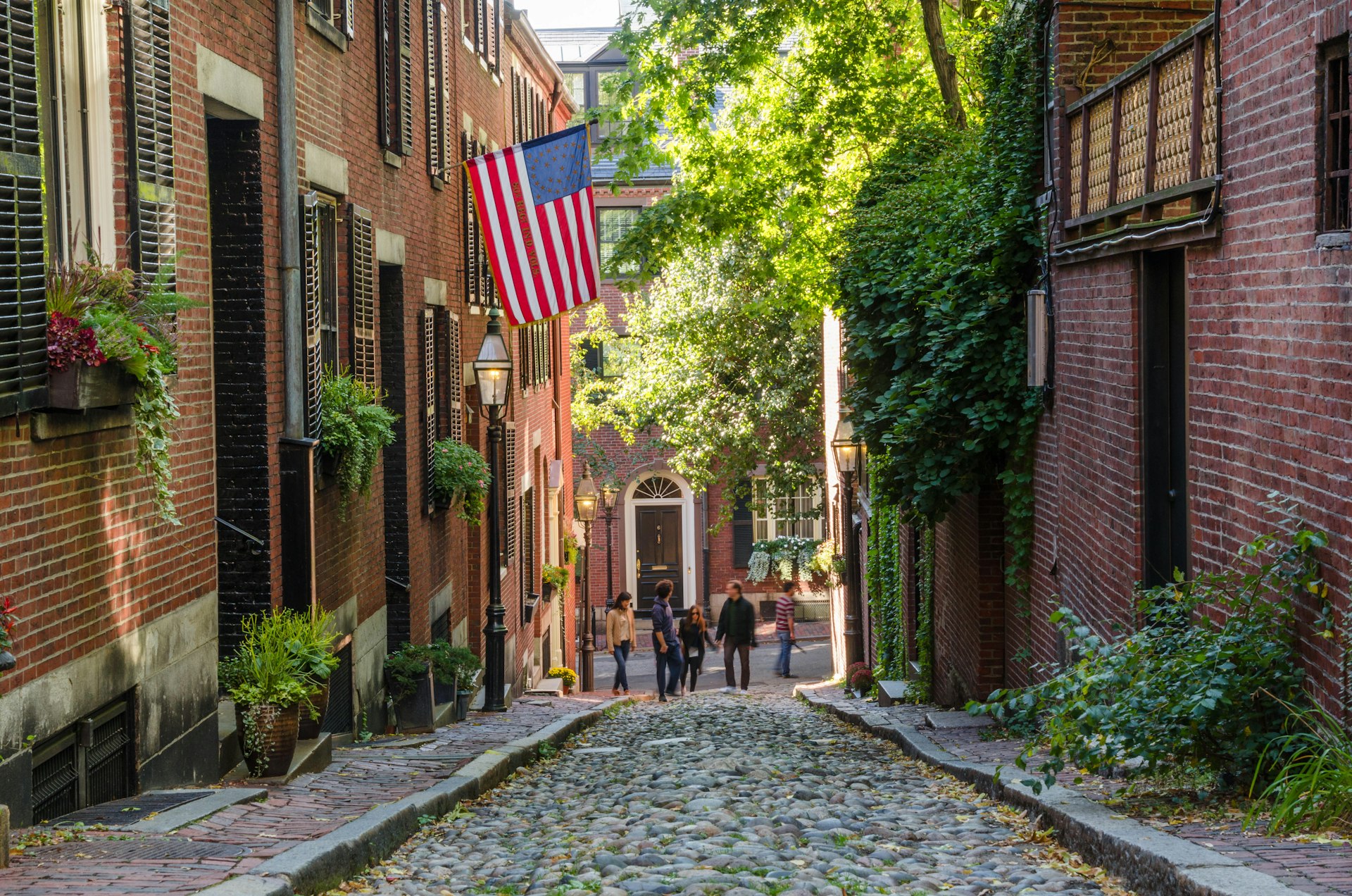 A narrow cobbled street lined with historic buildings. One has a large American flag flying outside it