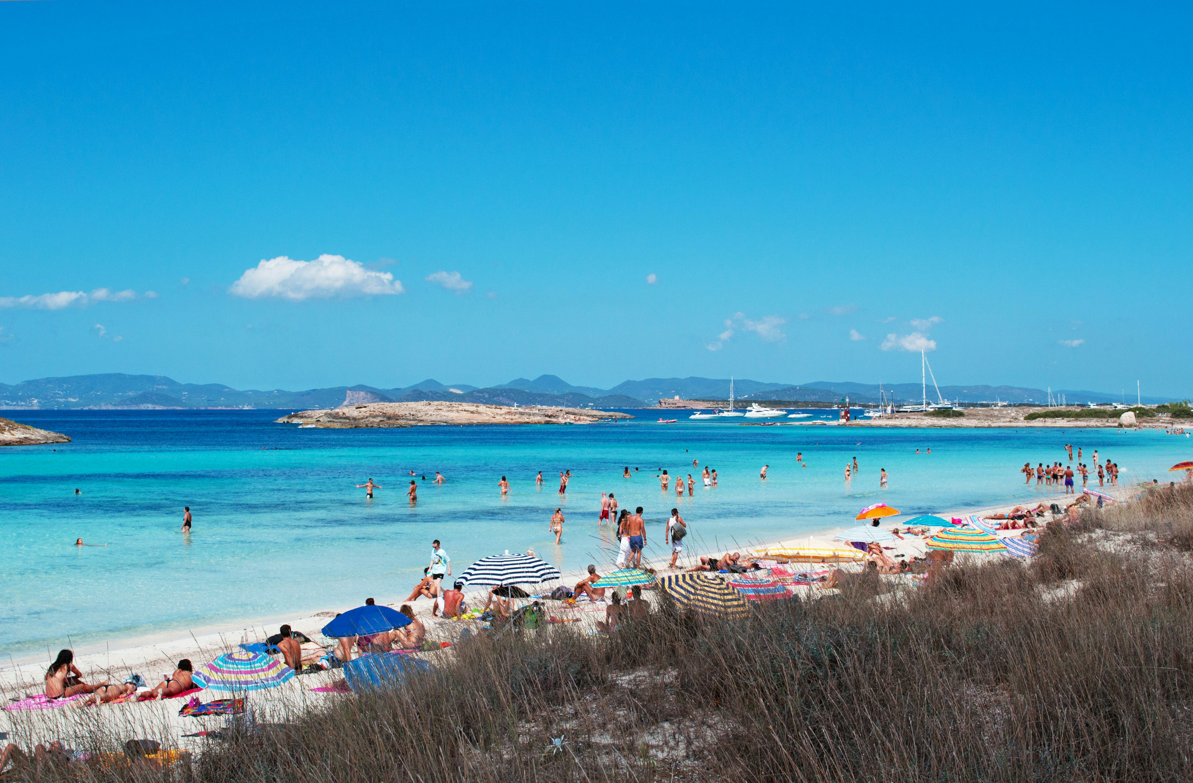 People relax on the beach and swim in the clear waters of a wide bay with two islets