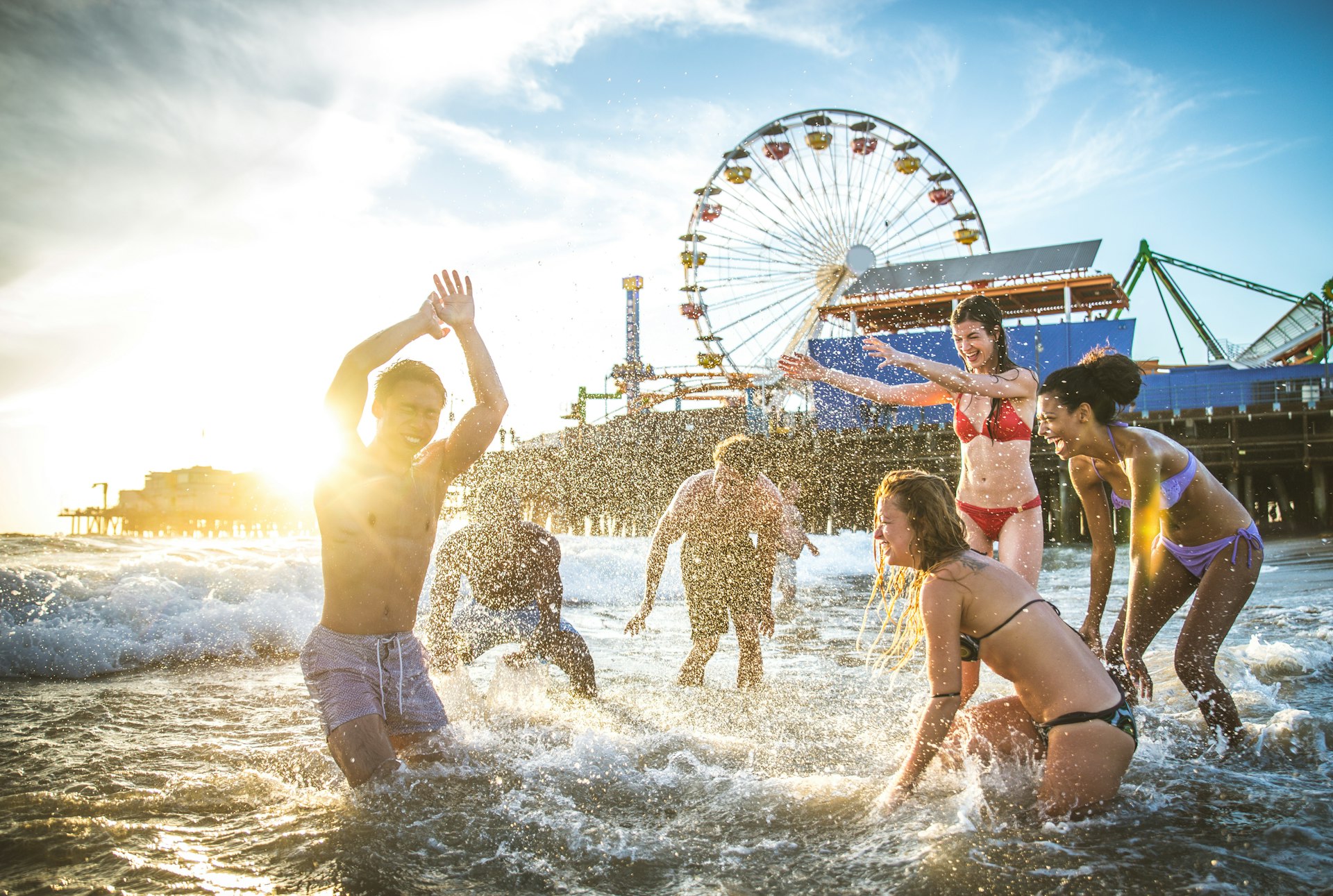 A group of friends splash in the shallow sea. Behind them is a large pier dominated by a huge Ferris wheel