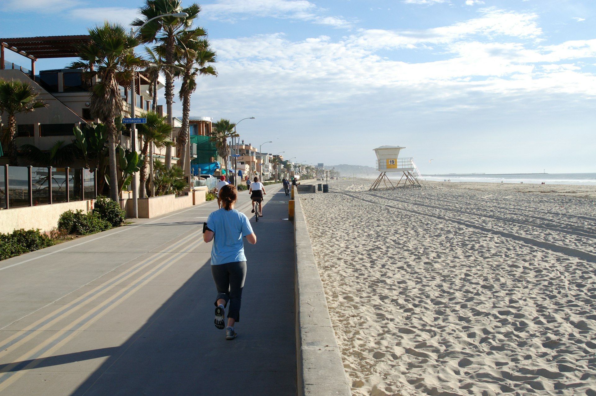 A jogger runs along Mission Beach Broadwalk in San Diego at sunrise with the golden sand of the beach to her right