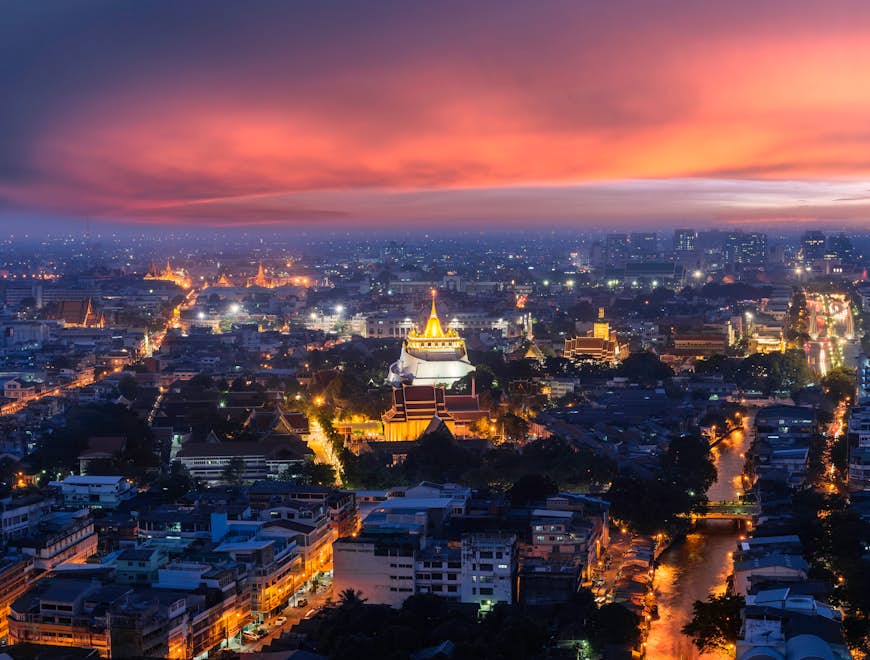 Wat Saket, the Golden Mount, shines brightly at dusk above the cityscape of Bangkok, which is drenched in a peach and orange sunset
