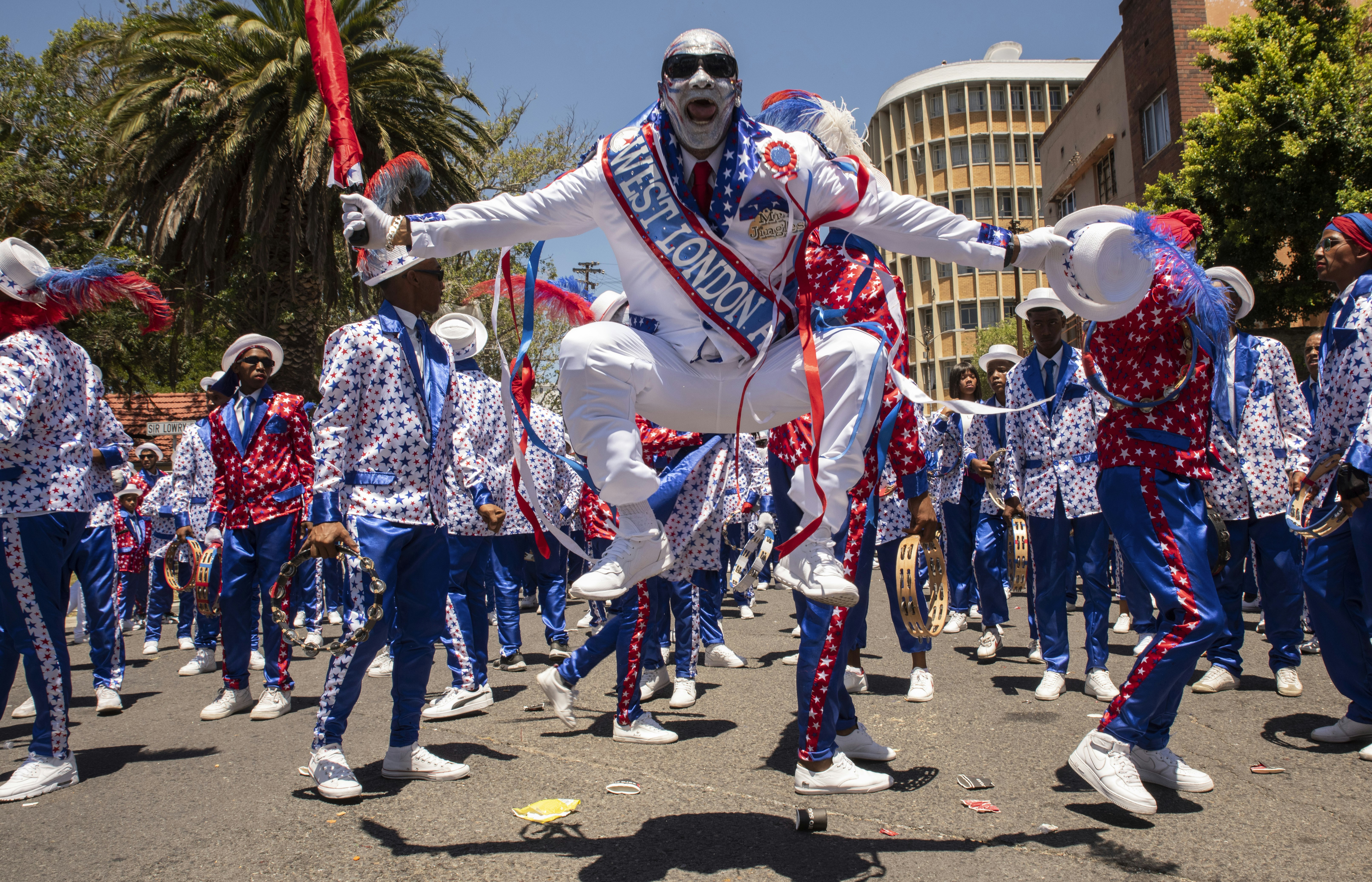 A man dressed in a white suit with a red, white and blue sash with the words "West London" on it jumps high in the air holding a white hat and red umbrella during the Cape Town Street Parade