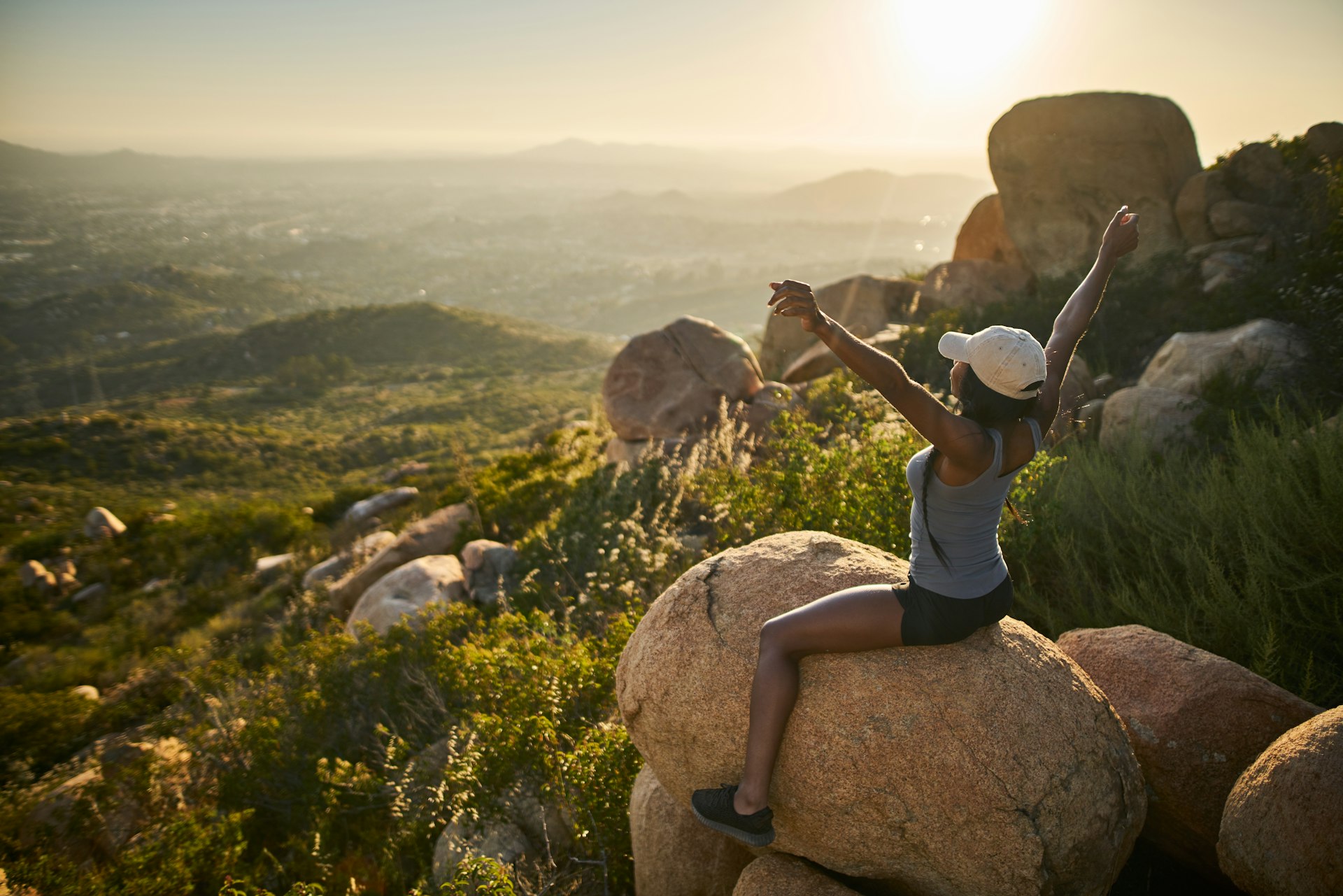 fit female hiker sitting on rock at mountain top looking at city in distance with arms up