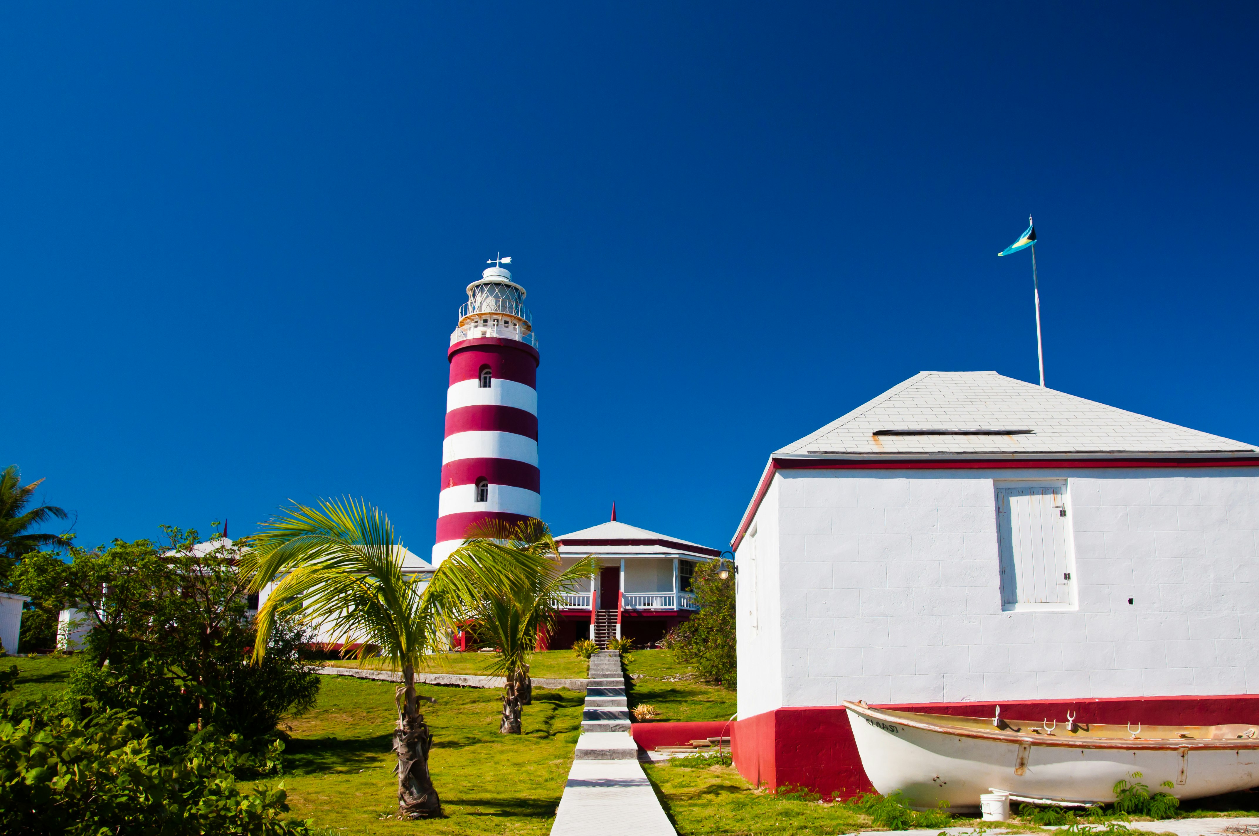 A paved path leads to a red and white house and candy cane stripped lighthouse on Elbow Cay in the Bahamas. Storage shed with small boat are in the foreground.