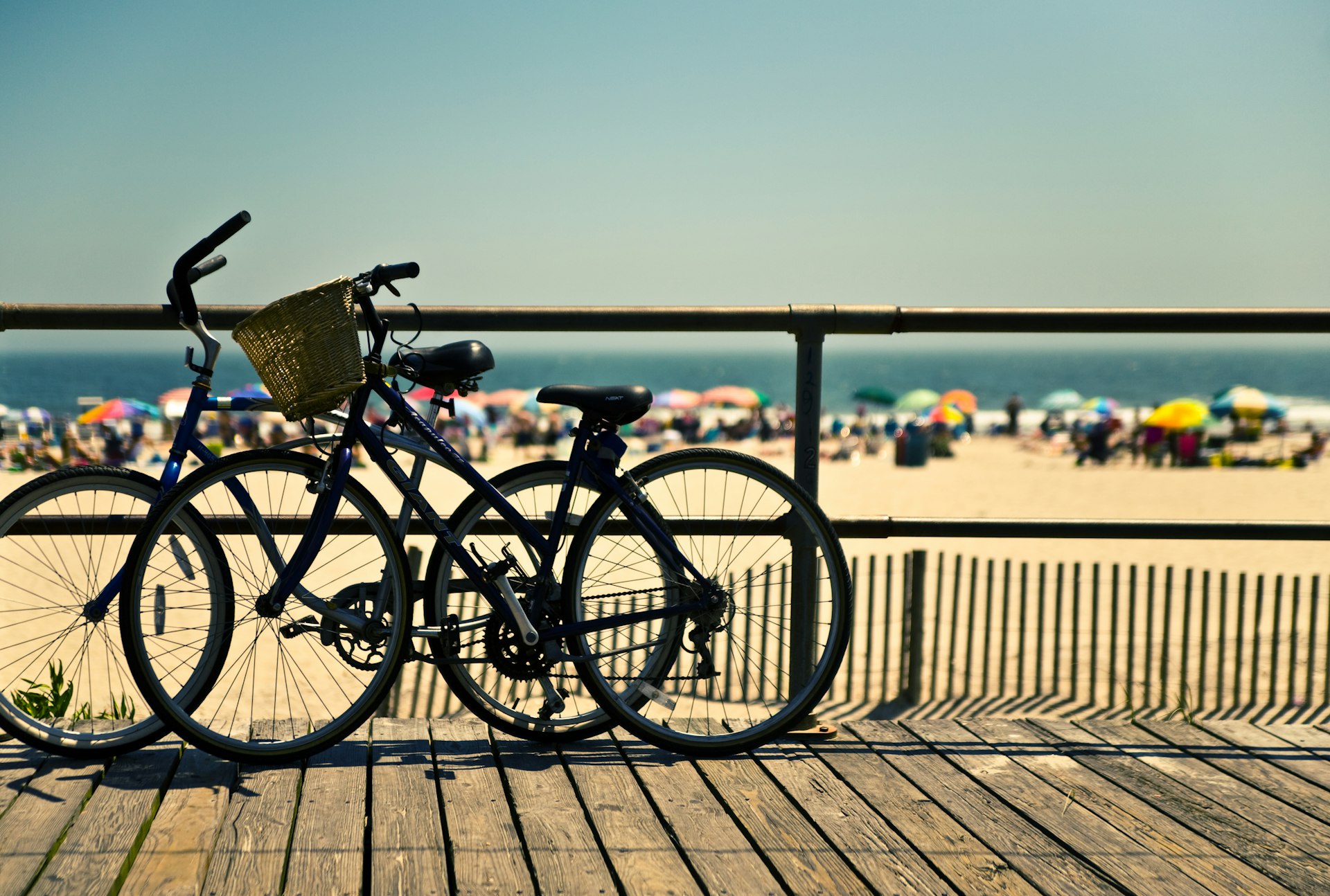 Bicycles in silhouette, propped by the edge of the boardwalk, against a backdrop of a crowded beach