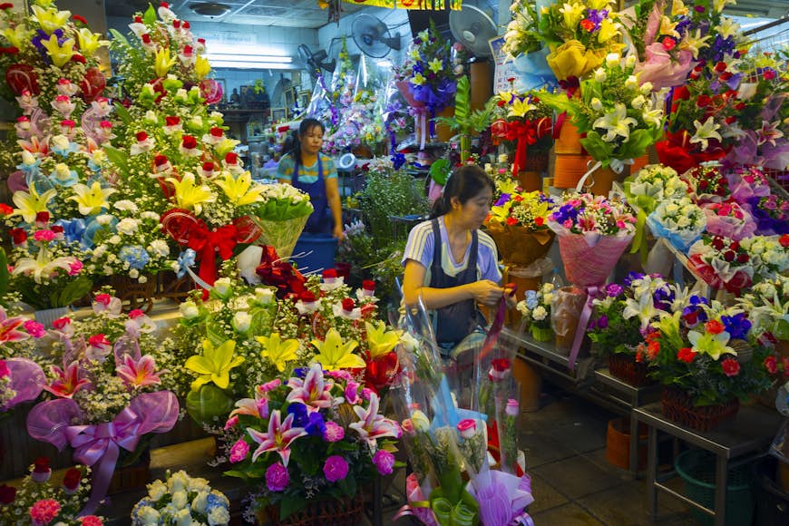 A female worker in a blue apron at work amongst the brightly-coloured flowers of the Pak Khlong Flower Market