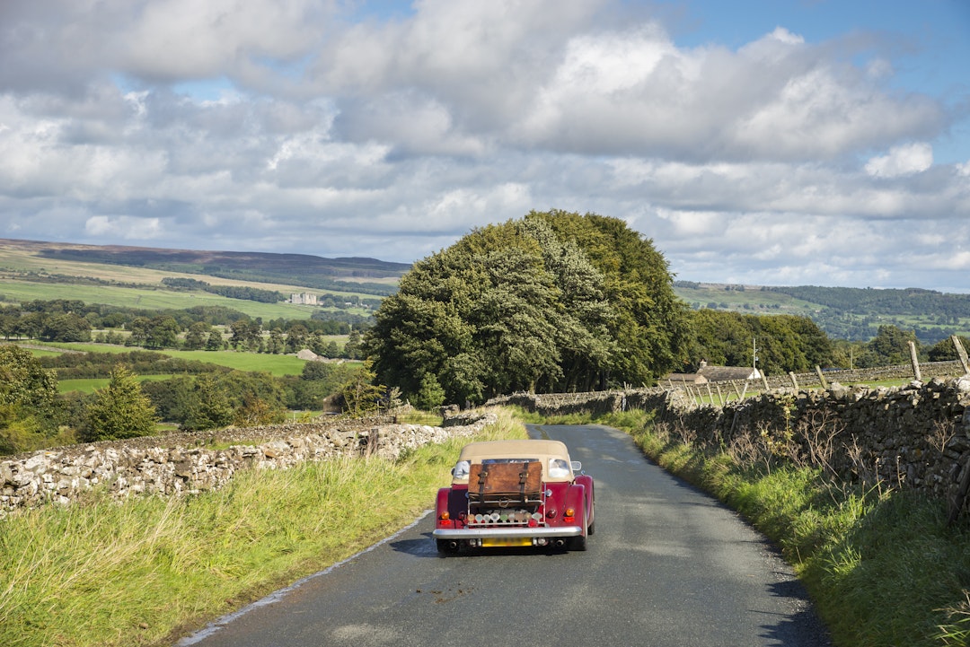Little red car driving near Aysgarth in the Yorkshire Dales on a sunny September day. Bolton castle seen in the far distance.