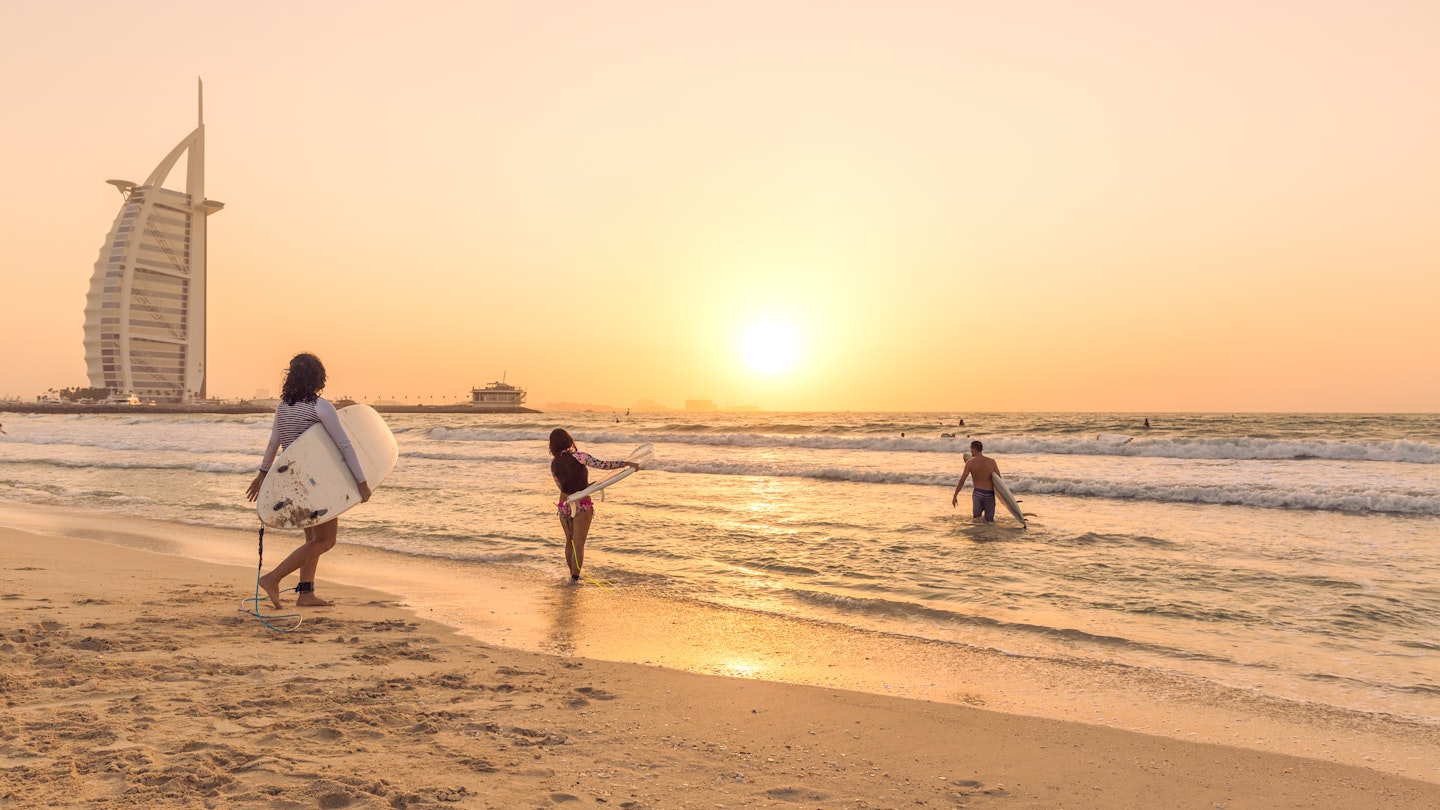 Dubai,UAE - October 04 2017: Three Surfers go into the sea from beach to catch a wave with Burj Al Arab hotel in background around sunset, Dubai, UAE.