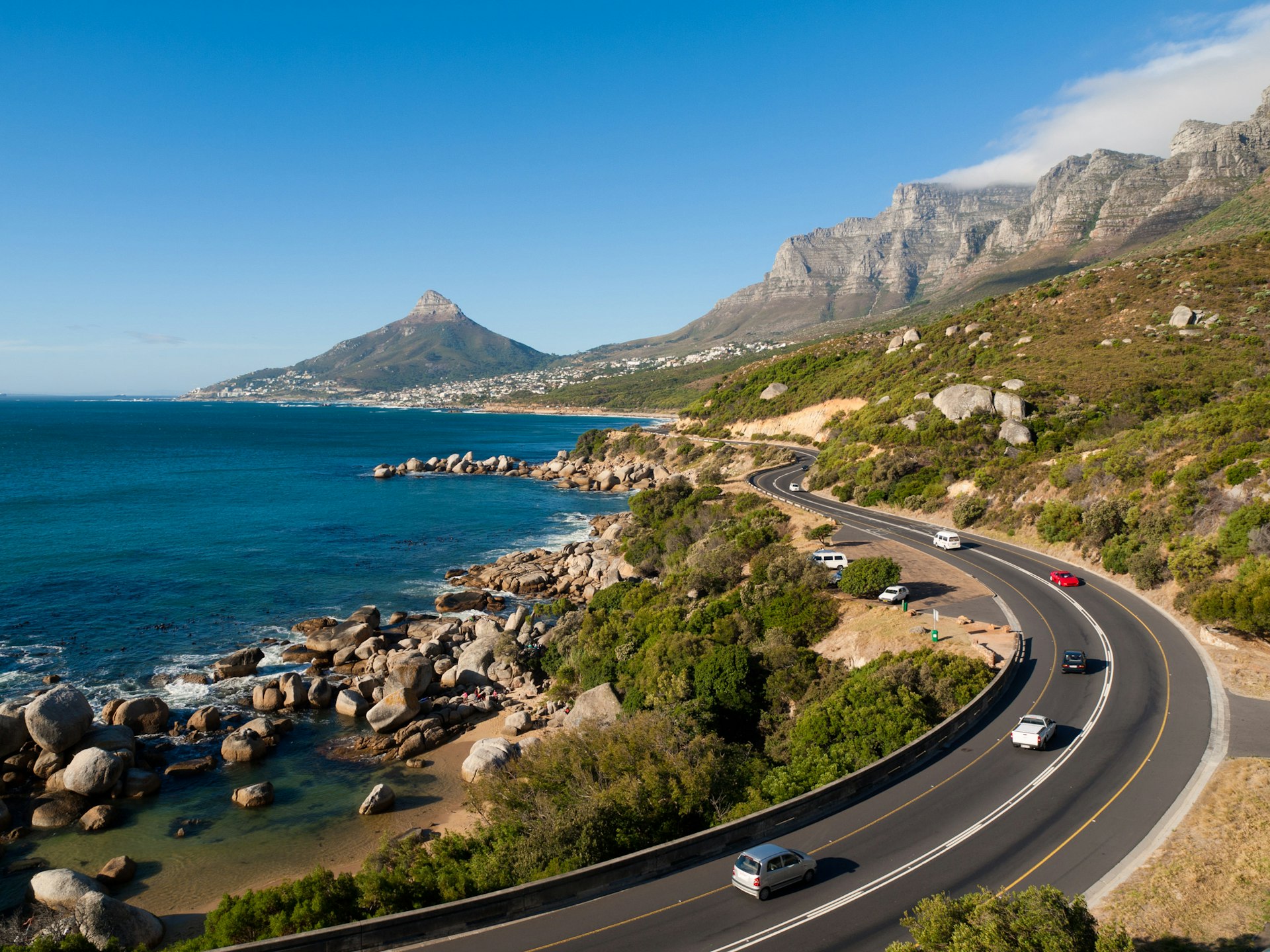 An aerial shot of a beach road, with distinct sandy covers. In the distance are rocky outcrops high to the right away from the sea