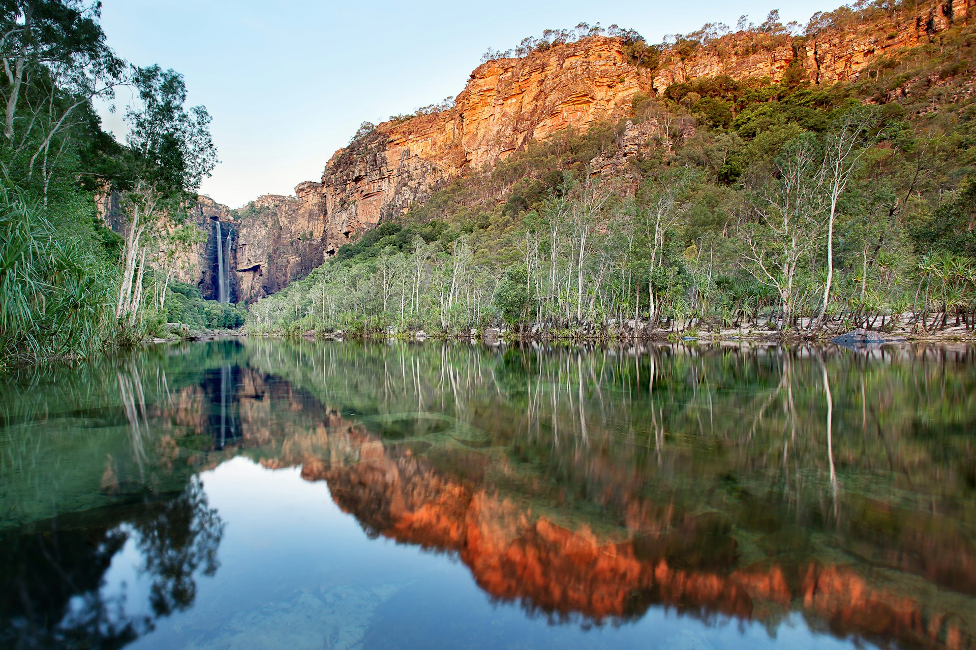 Glassy water is flanked by high trees and red cliffs on either side, with a waterfall at the furthest point from the camera. Kakadu National Park, Australia.