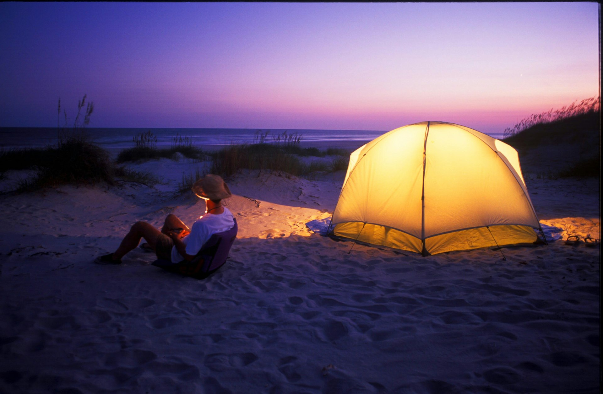 A man sitting on a low beach chair beside a glowing tent under the purple night sky at Hammocks Beach State Park 