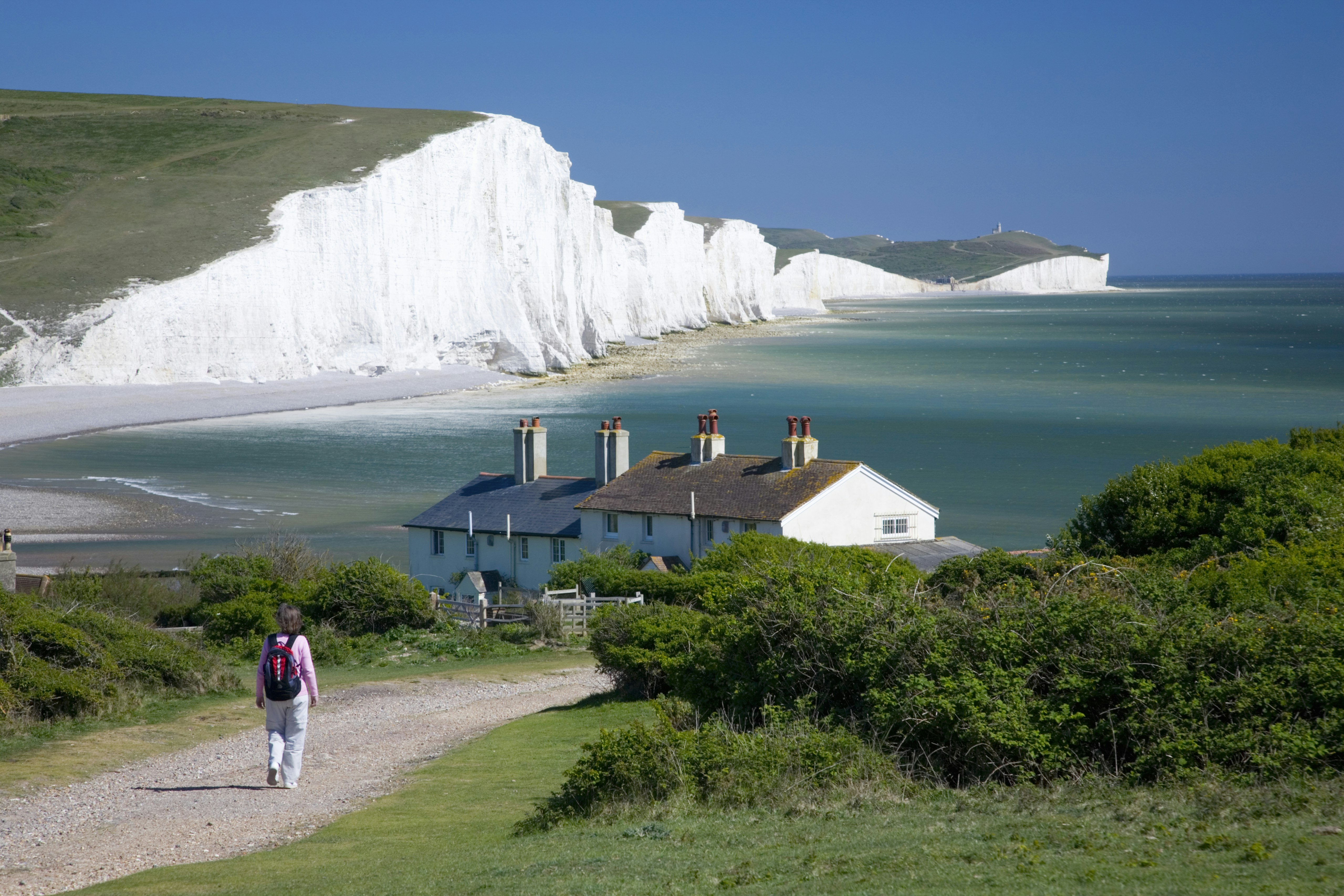 Person walking towards a small row of white cottages on a cliff. The white cliffs of Seven Sisters stretch out into the distance.