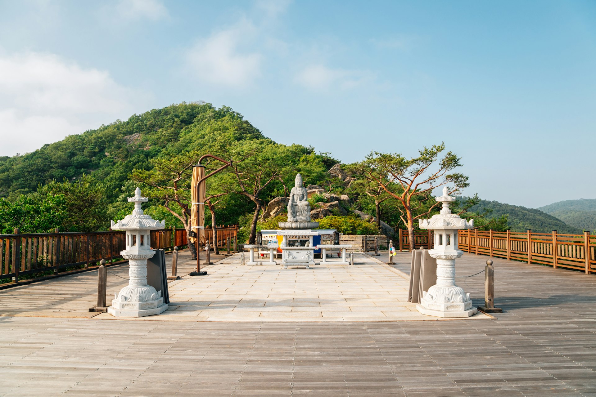 A statue of Buddha stands in a stone courtyard surrounded by hilltops that are covered in forest at Goryeosan mountain's Jeokseoksa temple in Ganghwado