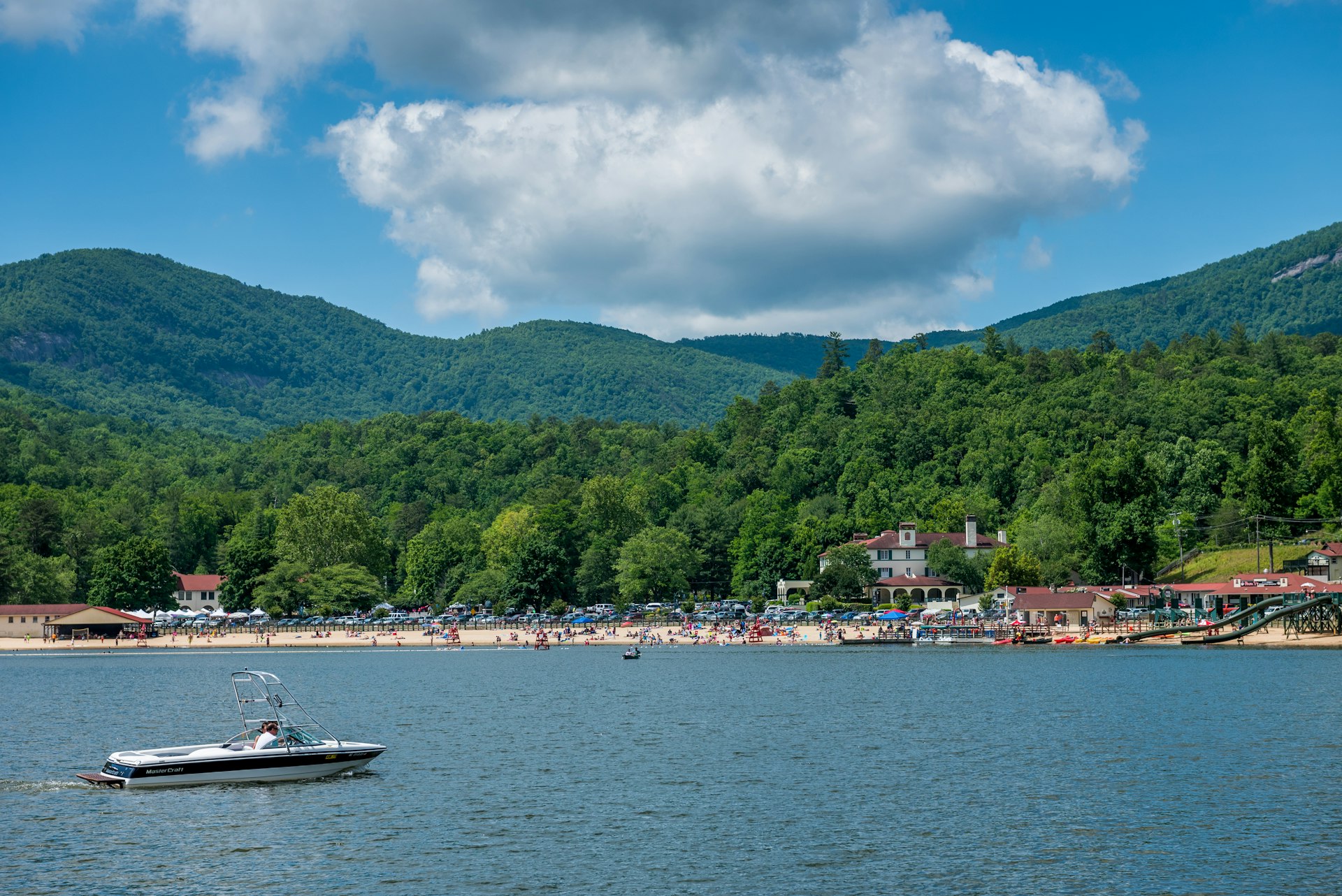 The beach at Lake Lure, North Carolina, in the early afternoon. People on the beach and boat in foreground. View of mountains in the background. 