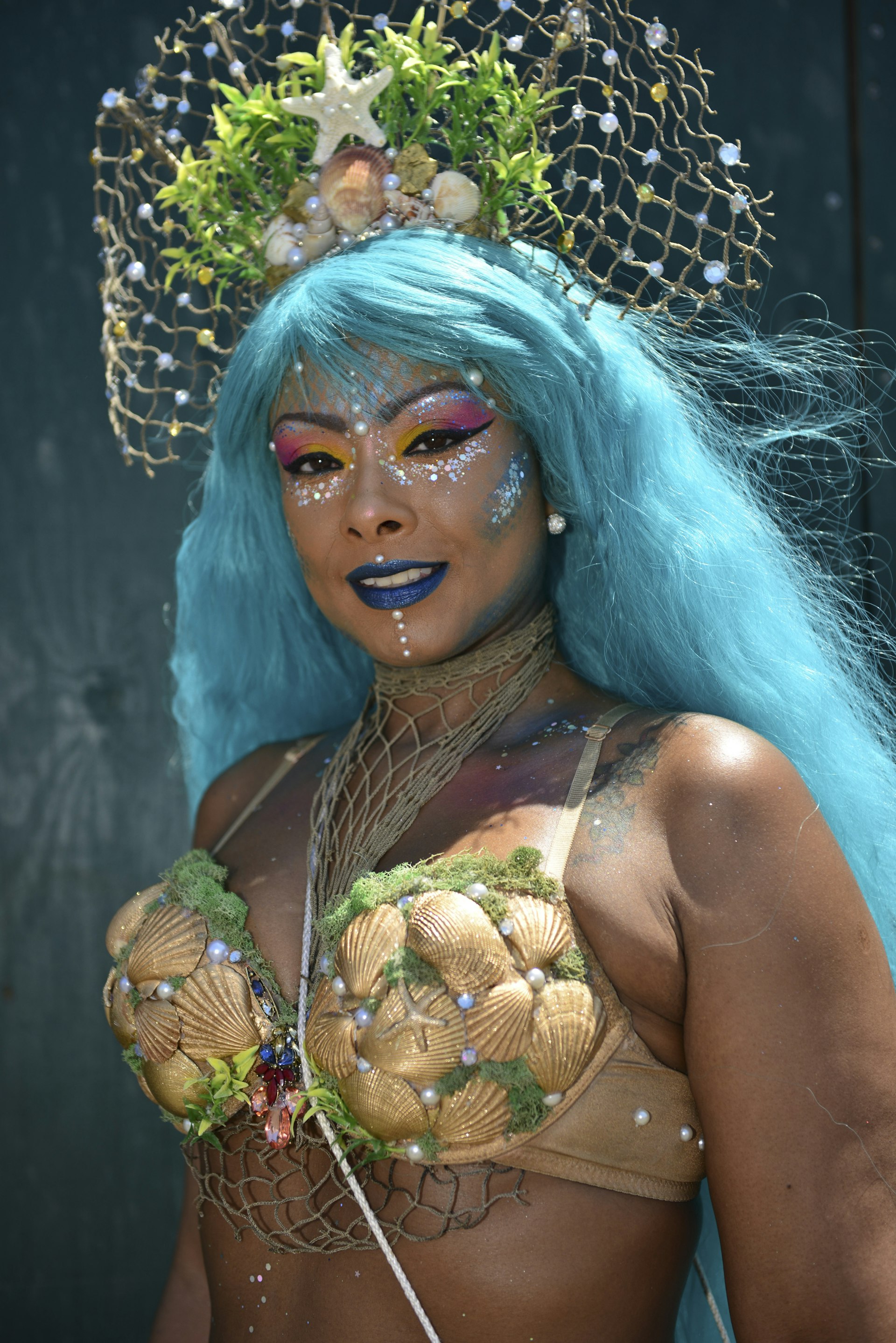 A person with an aqua-colored wig wears a golden crown filled with sea shells, a starfish and golden netting dotted with pearls. The person is wearing a bikini top made of sea shells that have been painted gold.