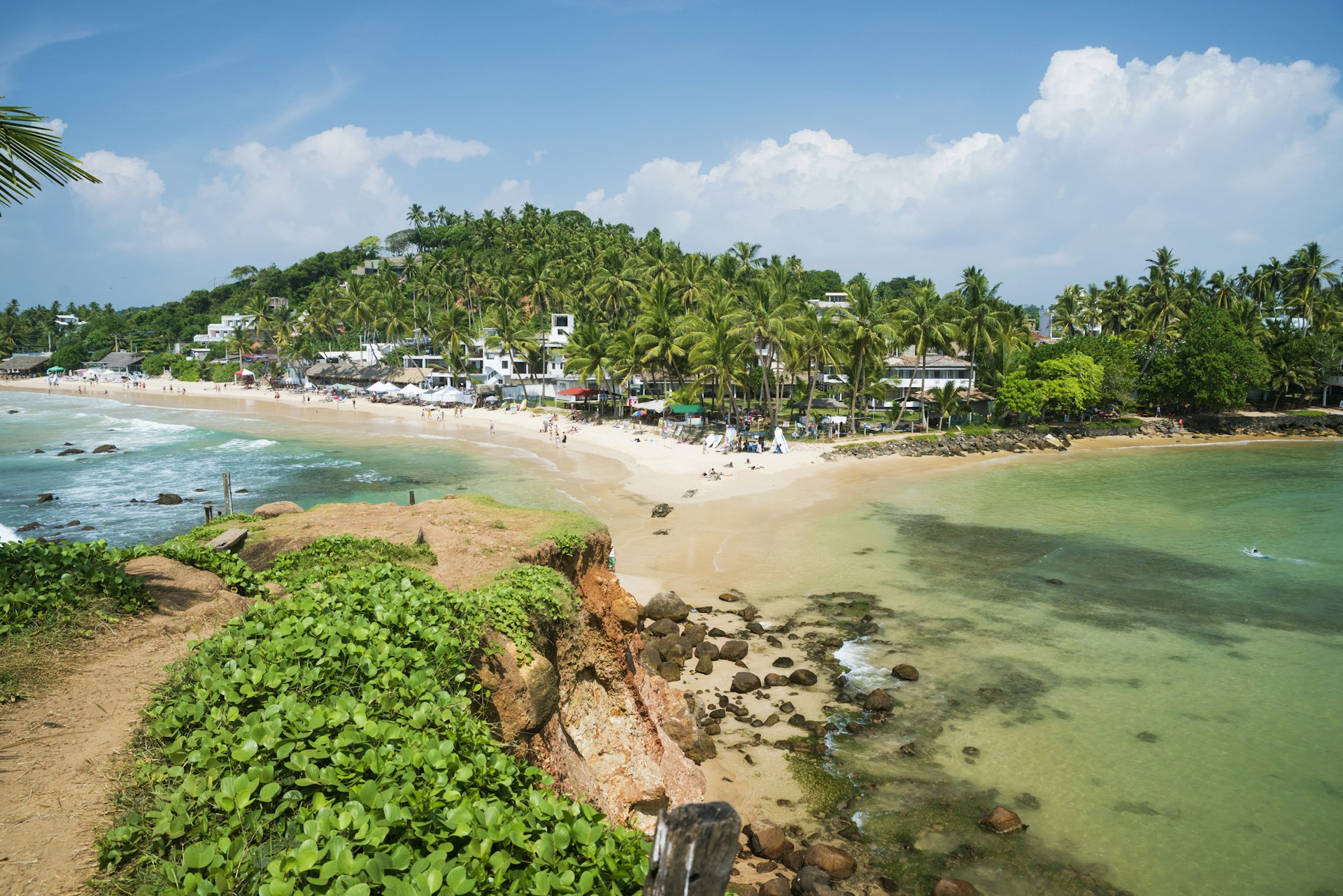 A view of the small beach resort of Mirissa on the south coast of Sri Lanka. The sandy, white beach curves out to a small islet, and is backed by palm trees.