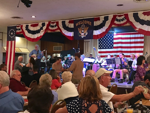 A stage decorated with the US's red-white-and-blue flag is full of seated musicians. In front of the stage two people are smiling and dancing