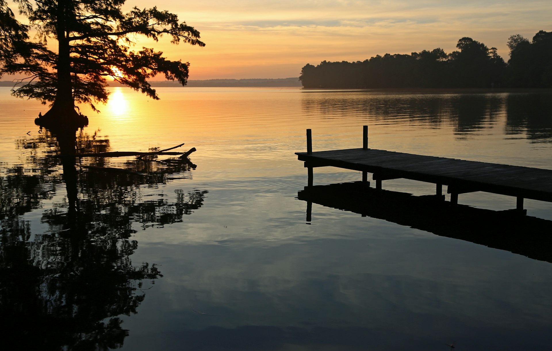The sun rises over Reelfoot Lake in Tennessee. There is a wooden pier jutting into the lake and trees dotting the shores. 