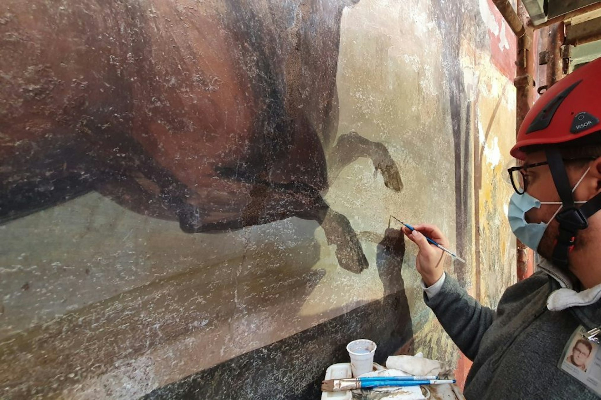 A person in a helmet and mask holds a paintbrush as they work to restore a historic fresco. 