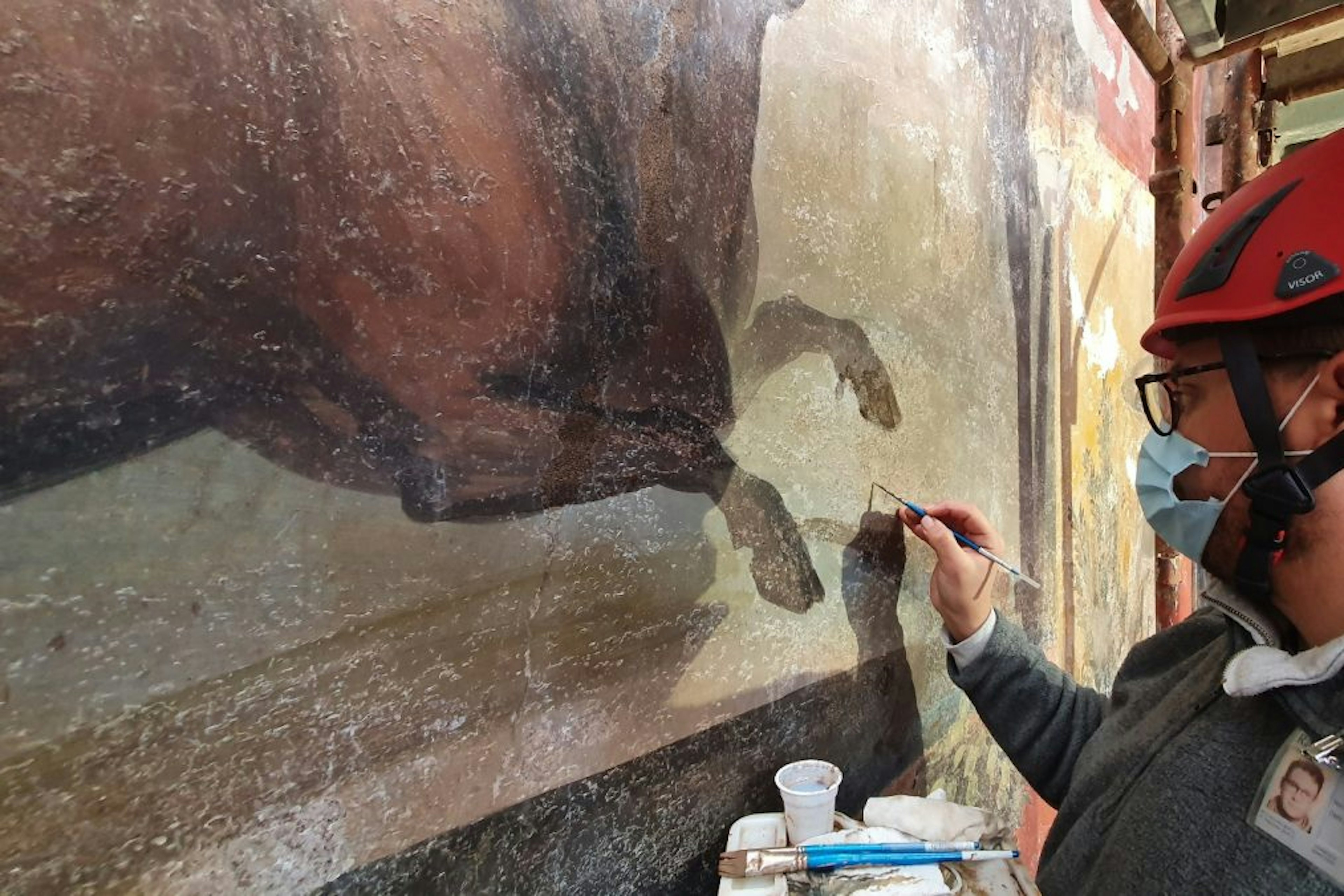 A person in a helmet and mask holds a paintbrush as they work to restore a historic fresco.